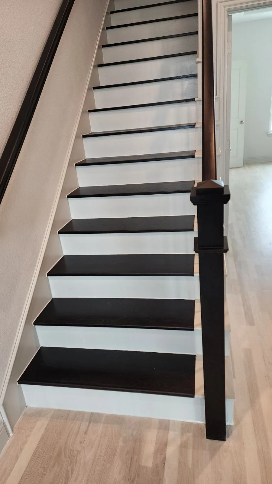 Inside view of a staircase with black wooden treads and white risers, black handrail on the right side, leading to an upper floor, with a door visible in the background.