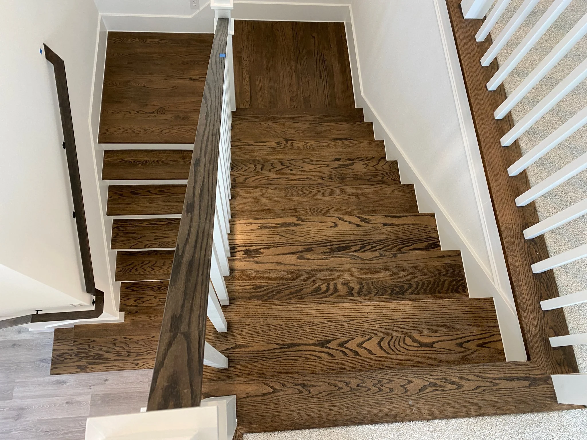 Wood staircase viewed from above with white risers, dark handrail, and wooden flooring at the top and bottom of the stairs, surrounded by white walls.