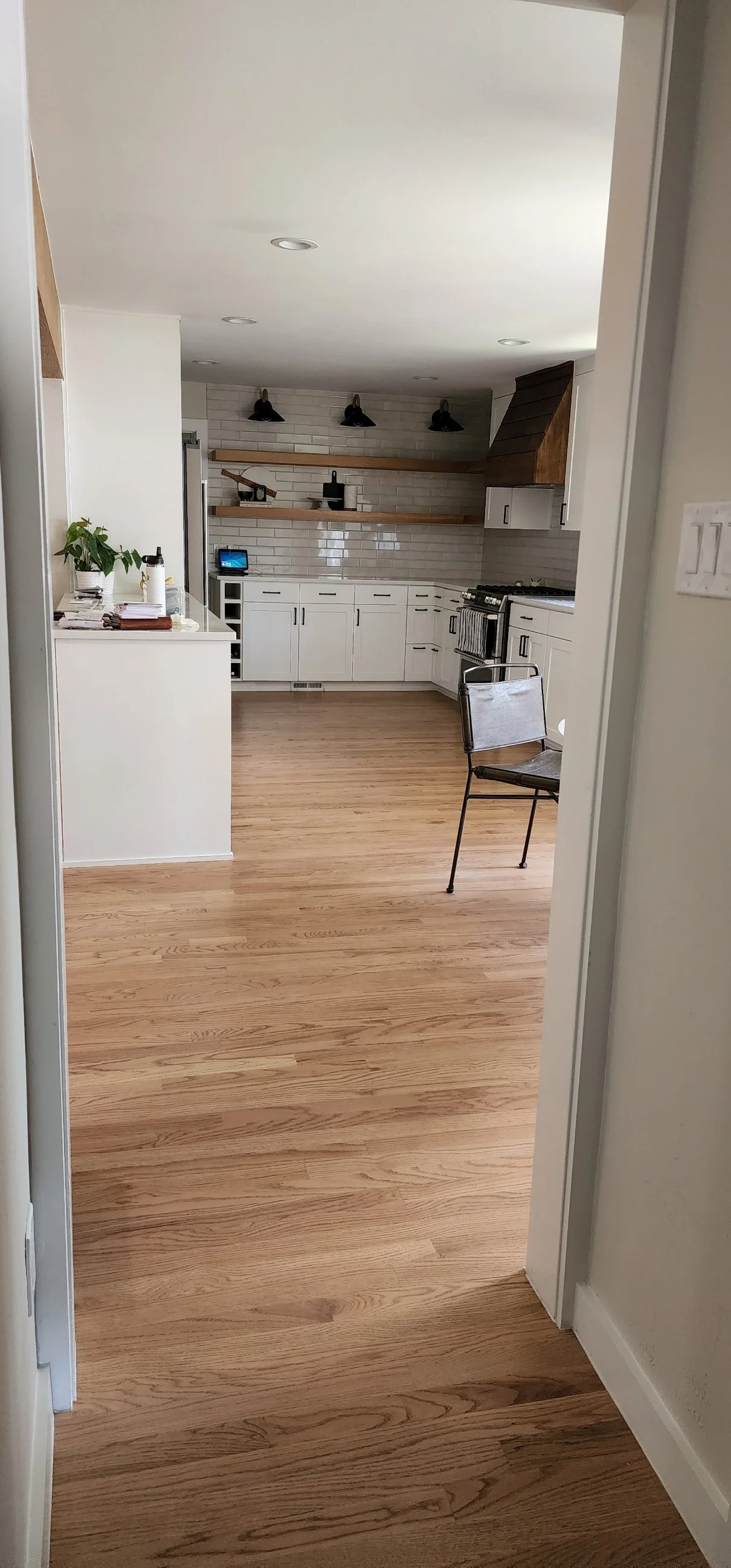A modern kitchen with white cabinets, wooden open shelves, black light fixtures, a stainless steel oven and stove, and light wood flooring. There is a small desk with a plant and some objects on it, and a metal chair with a cushion.