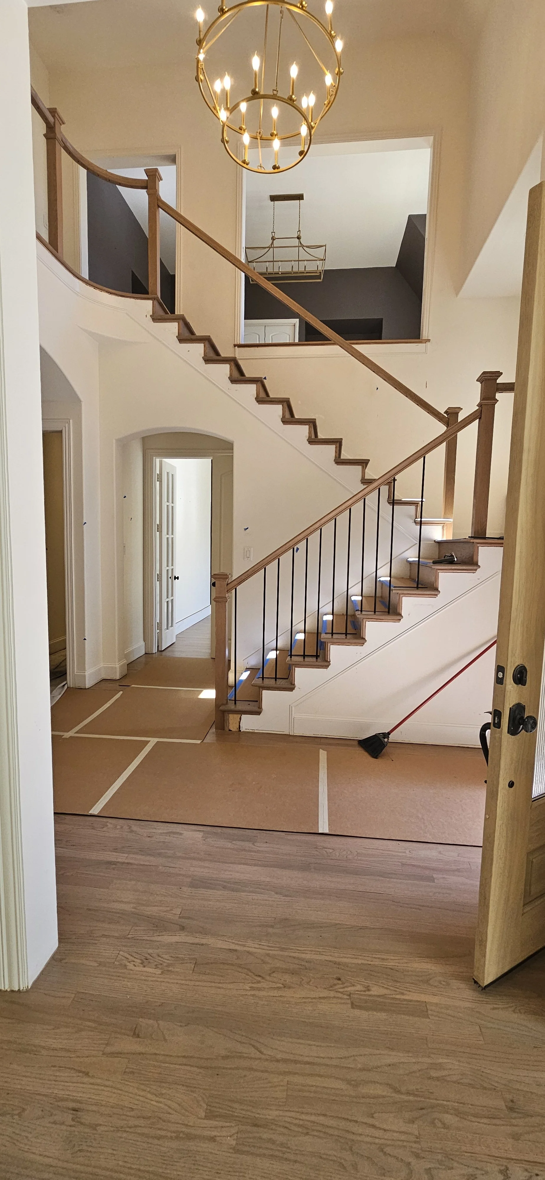 An interior view of a house showing a staircase with wooden handrails and black balusters, chandelier lighting, and a partially visible doorway. The floor appears to be in the process of renovation with protective covering and a broom visible.