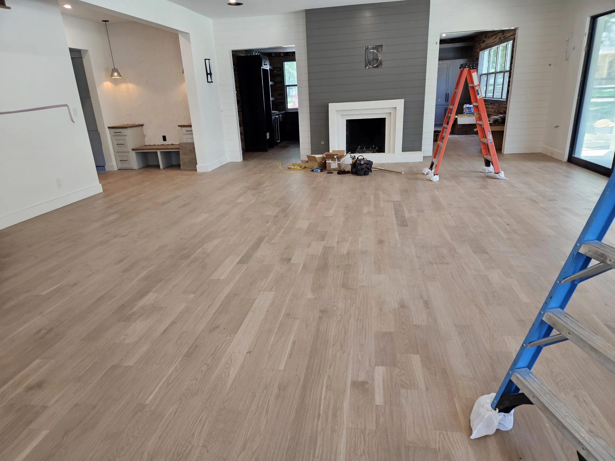 Living room under renovation with new hardwood flooring, ladders, tools, and construction materials visible near the fireplace.