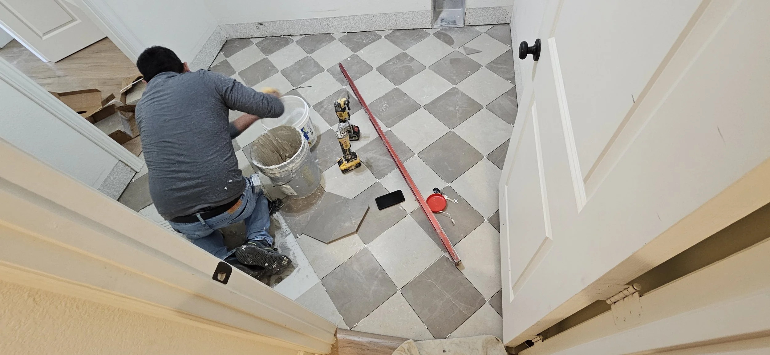 A person kneeling on a tiled floor, working on installing or repairing tiles, with tools and construction materials around.