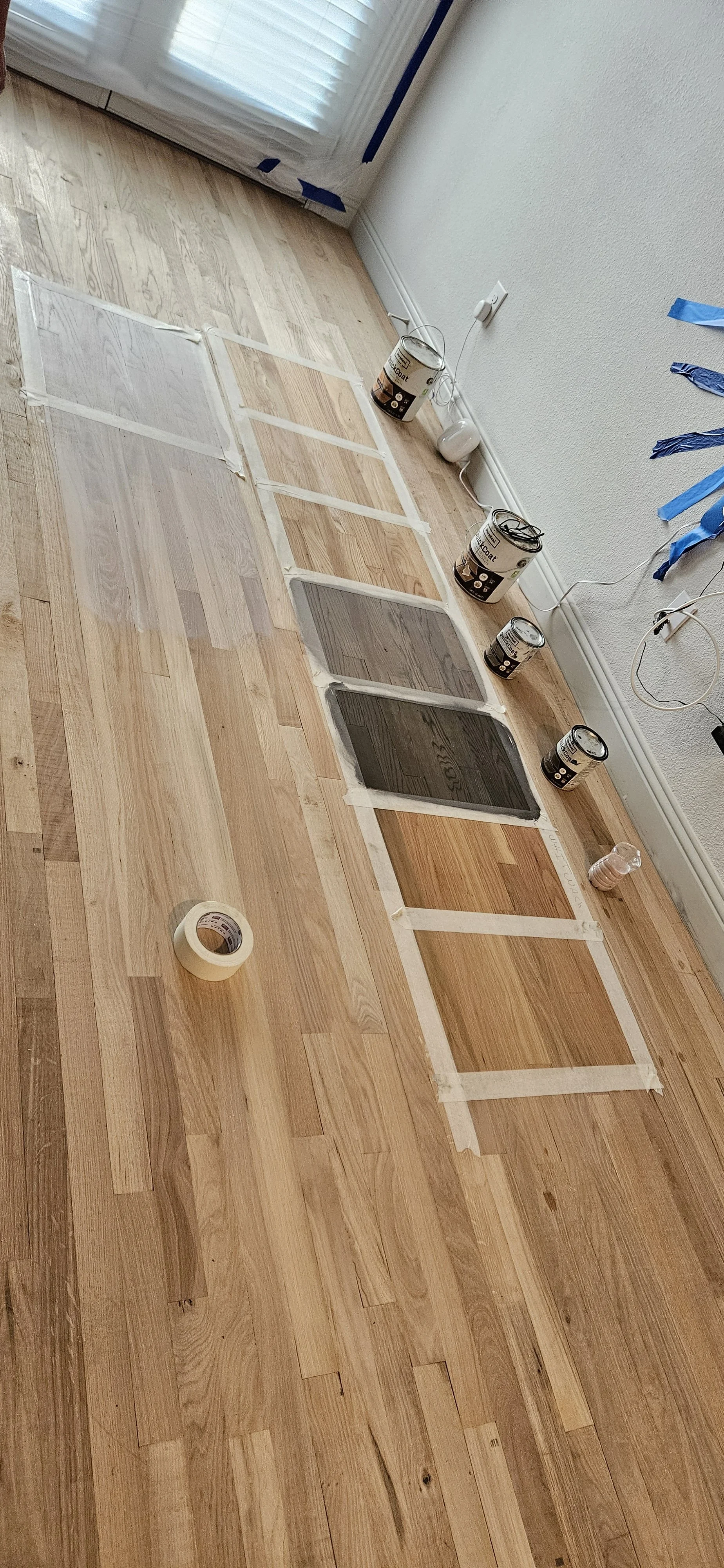 Freshly sanded hardwood floor with darker stain samples on taped paper, paint cans, a roll of painter's tape, a water bottle, and a partially taped wall near a window with blinds.