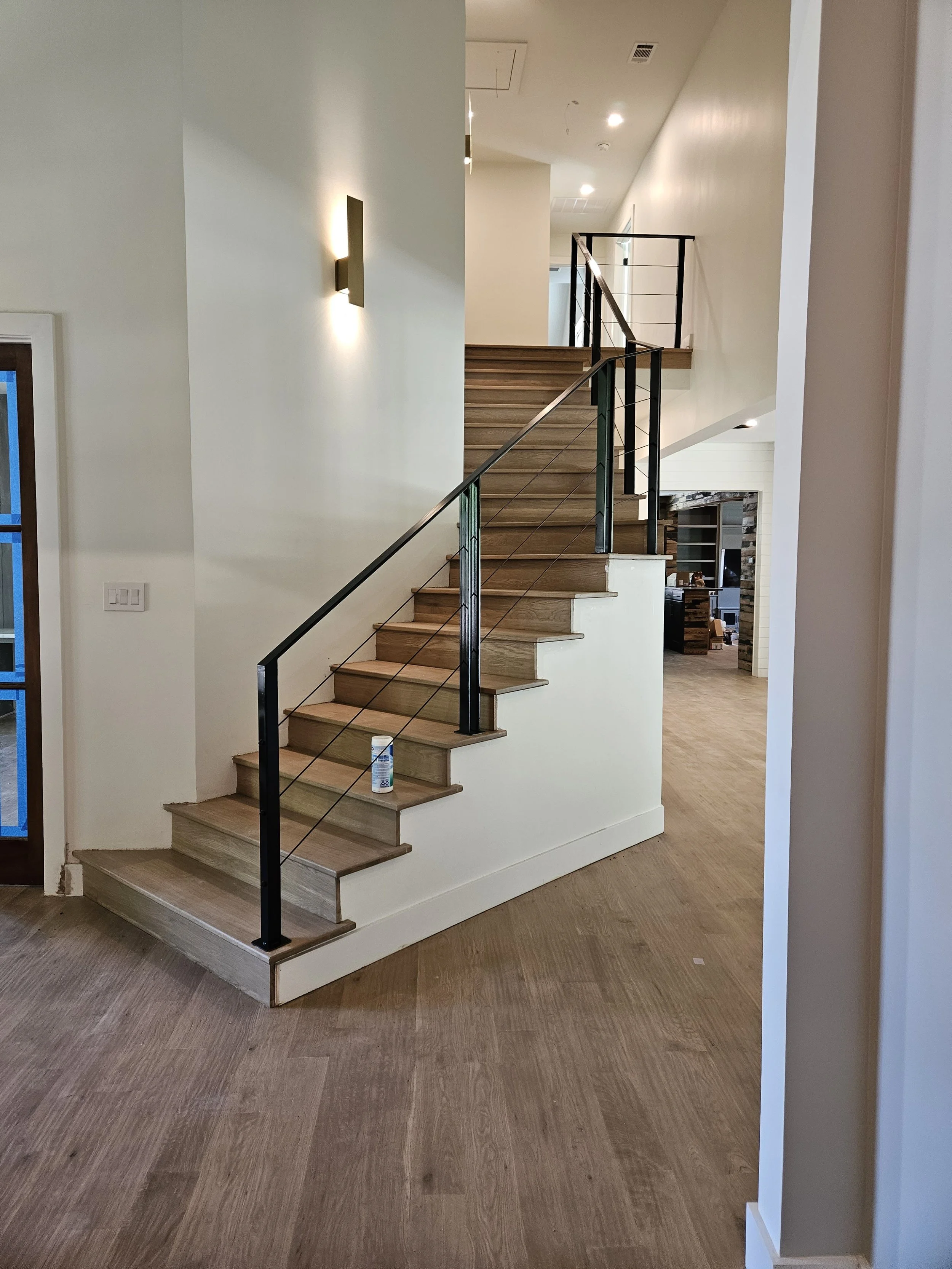 Interior view of a modern home showing wooden stairs with a black metal railing, white walls, and hardwood floors. There is a wall-mounted light fixture and a can of spray near the bottom of the stairs.