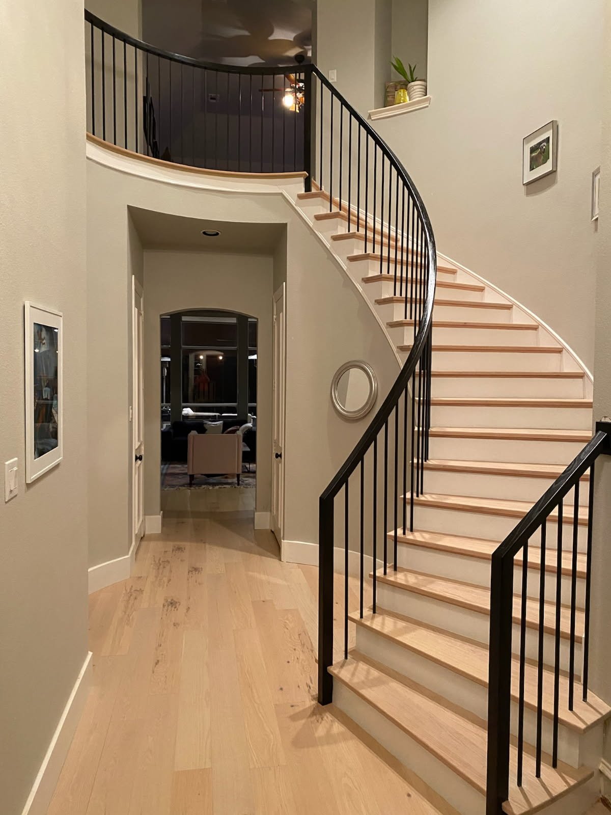 An interior view of a house entryway with a curved staircase. The staircase has light wooden steps and black metal railings. The floor is also light wood. There are framed pictures on the walls and a small circular mirror. In the background, there is