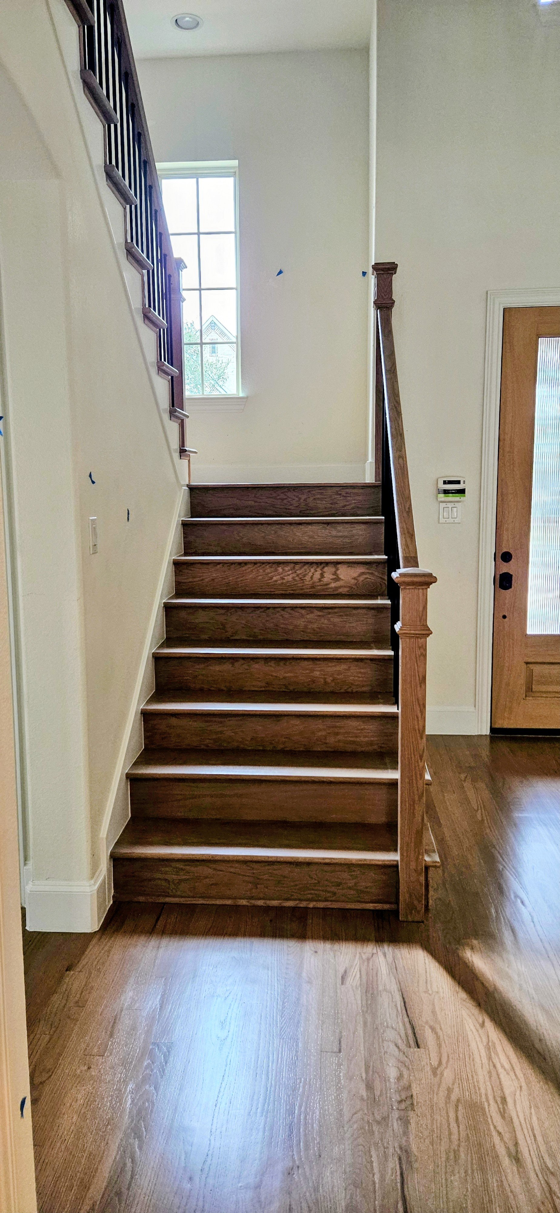 Interior view of a wooden staircase leading to an upper floor, with a window in the background letting in natural light. The stairs and flooring are made of dark wood, and there is a front door with a glass panel on the right side.