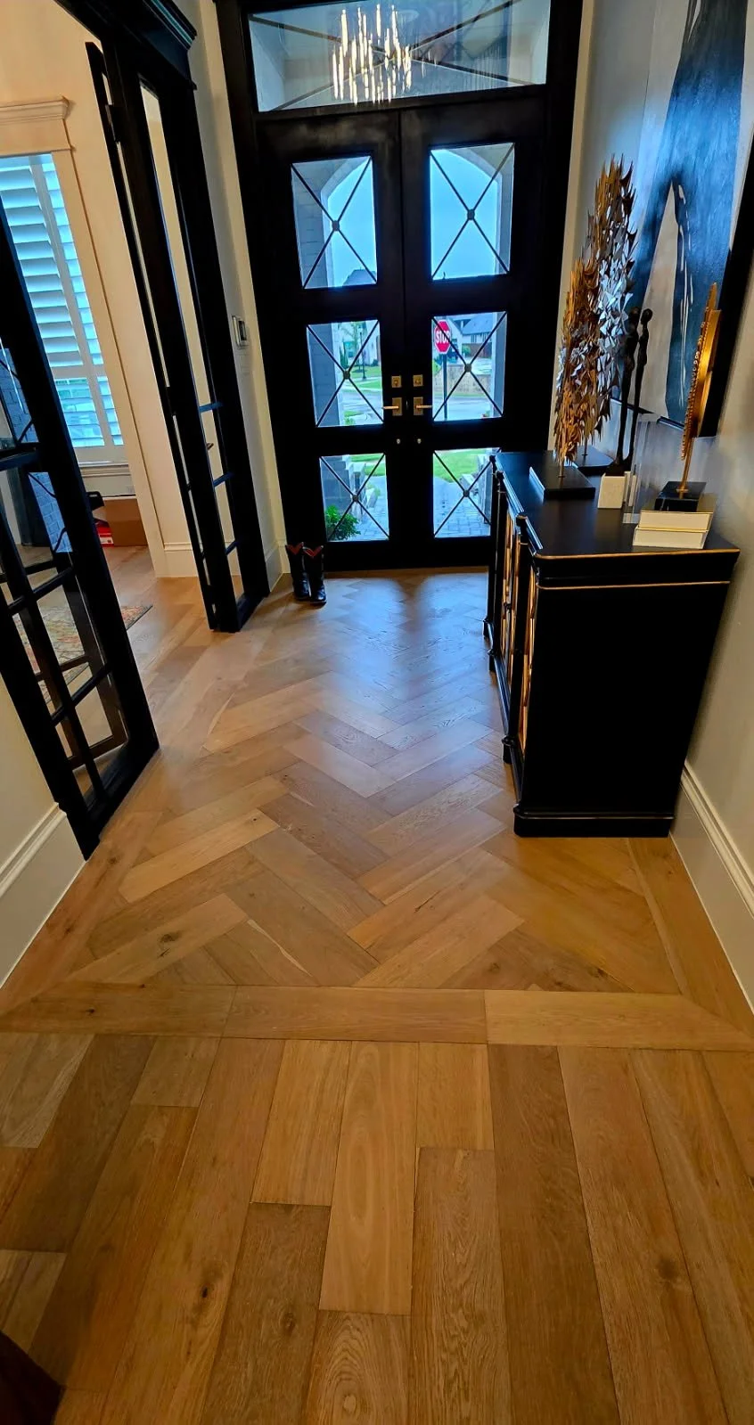 Entryway with a black front door, hardwood floor with a chevron pattern, and a black cabinet with decorative objects. Natural light coming through glass panes in the door and window on the left.
