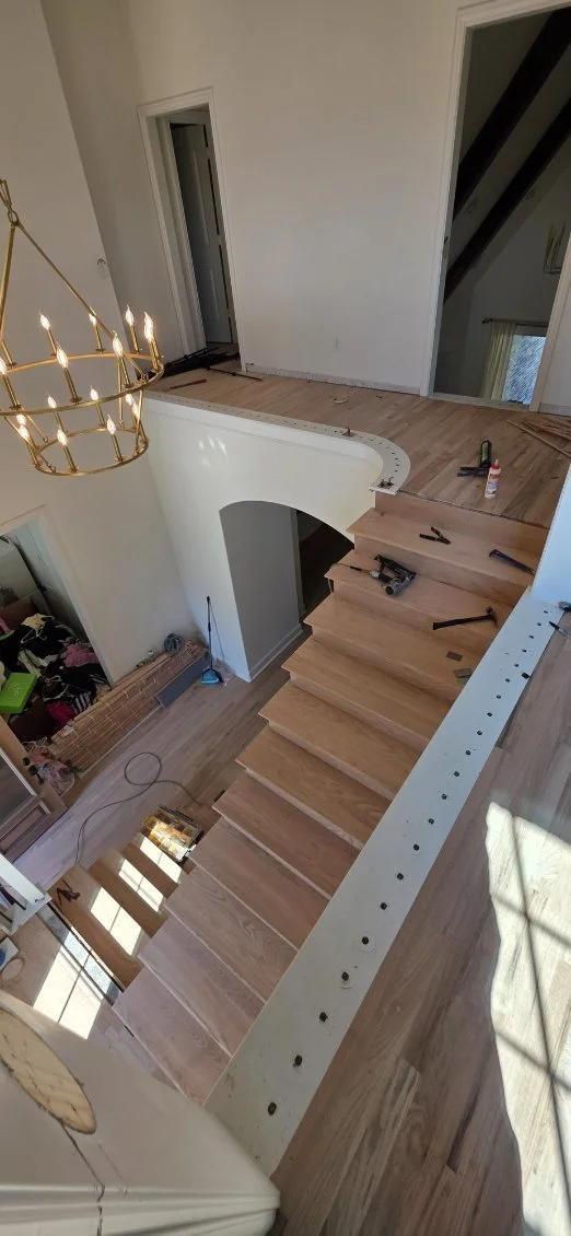 A staircase under construction in a home interior, with wooden steps and tools lying nearby. The photo is taken from an upper floor looking down into the foyer, with a chandelier hanging above.