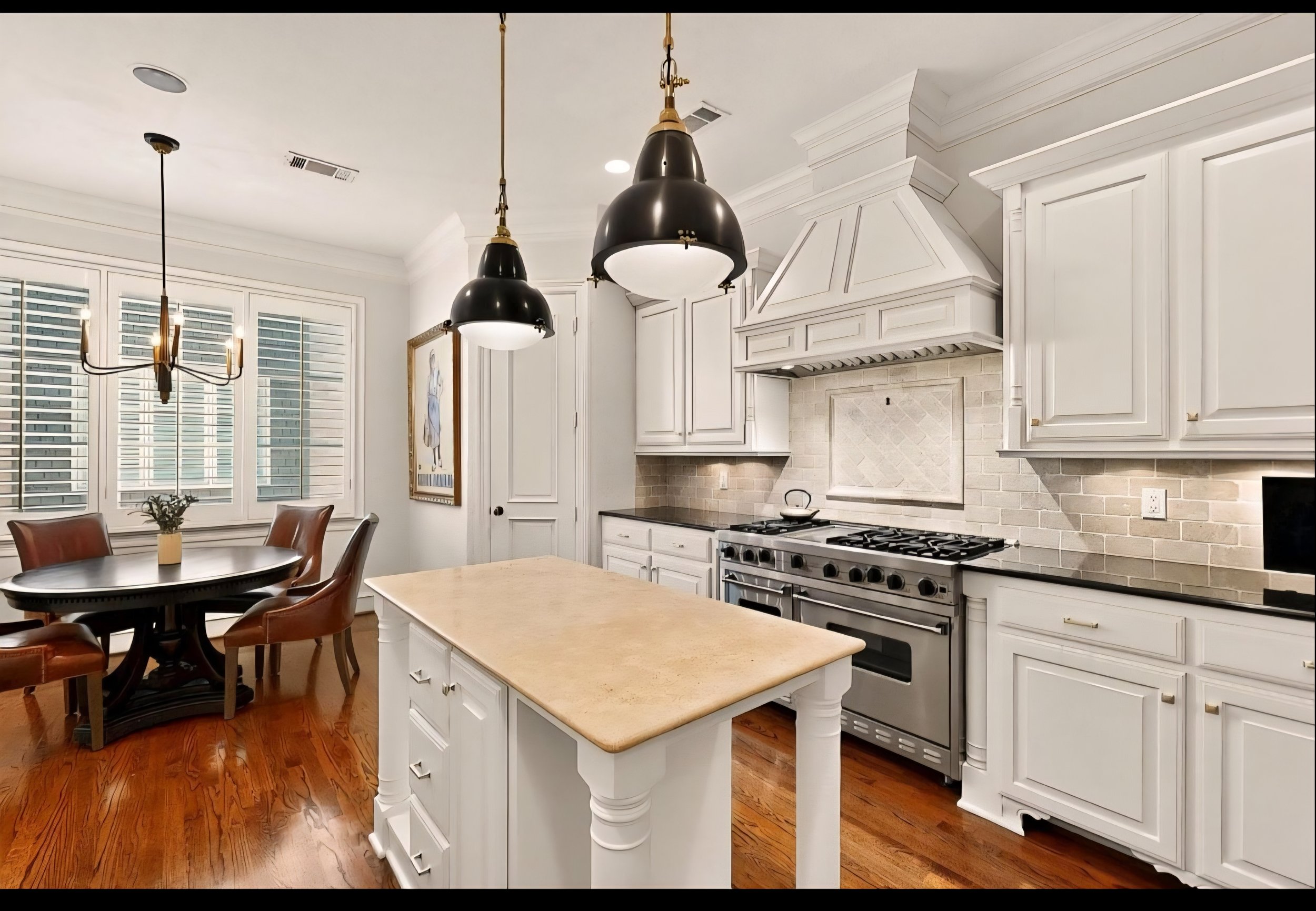 A modern kitchen with white cabinetry, a beige countertop island, a stainless steel oven and stove, black and brass pendant lights, a dining area with a round table and four brown leather chairs, a framed artwork, large windows with white shutters, a
