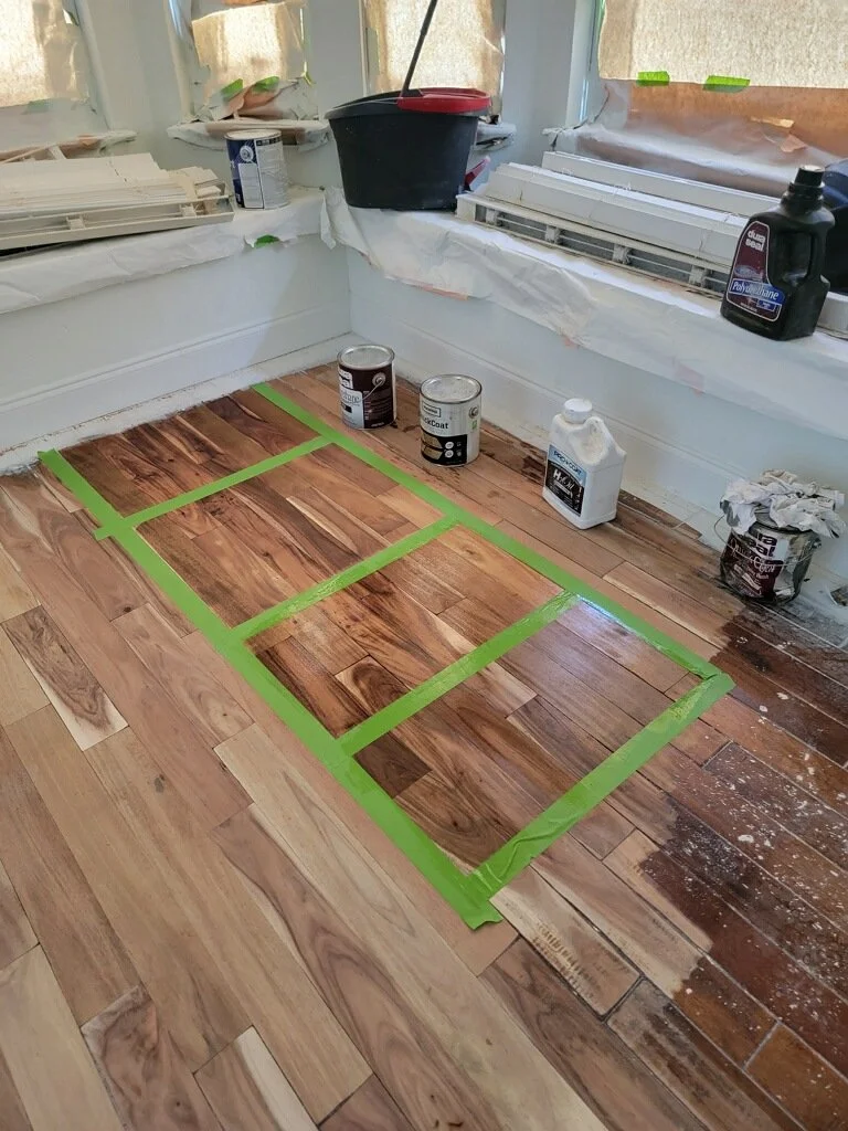 Room with hardwood flooring under renovation, partially sanded and stained, with green painter's tape outlining sections for staining or finishing. Painting supplies and equipment are visible near the windows.