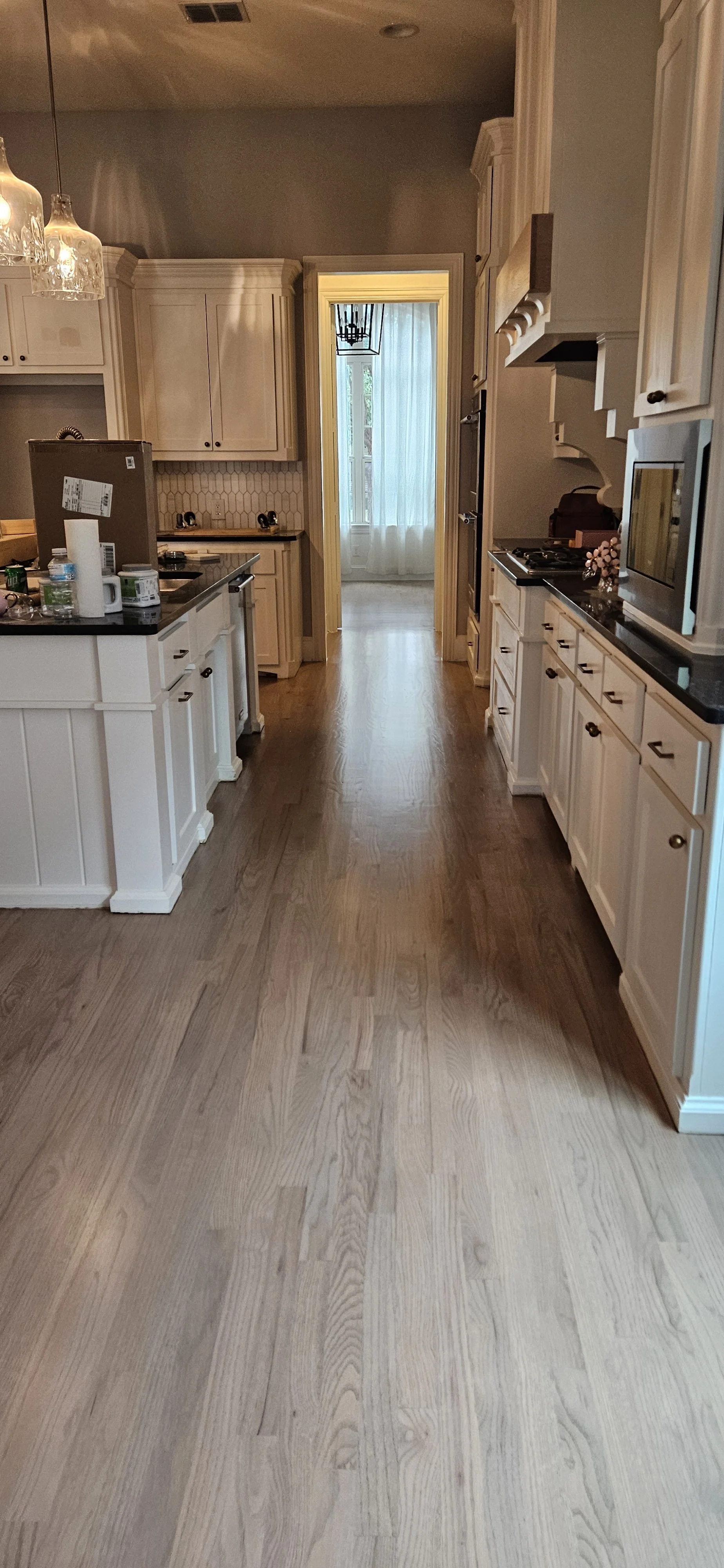 A view of a kitchen with white cabinets and dark countertops, hardwood floors, and a doorway leading to a sunlit room with curtains and a chandelier.
