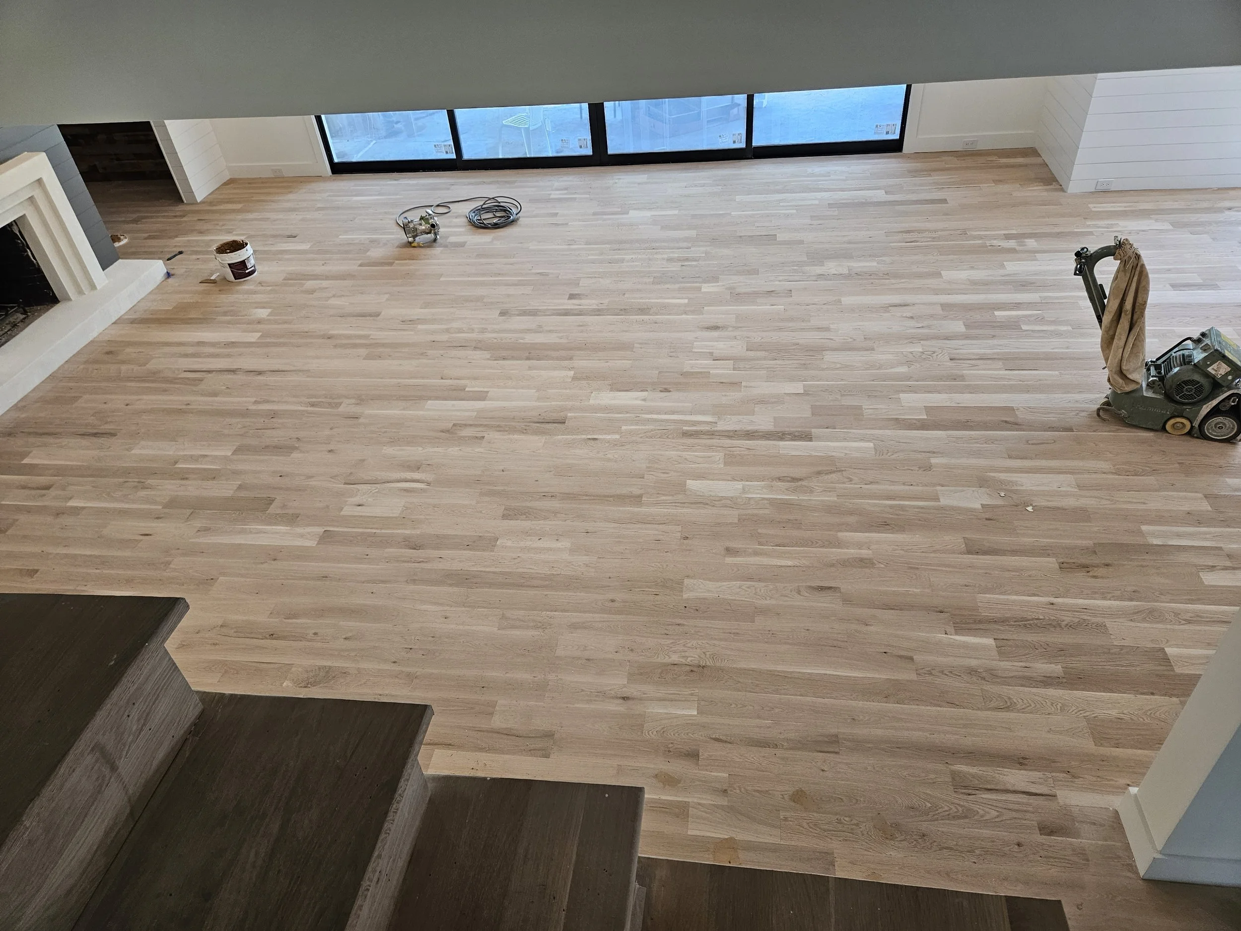 Empty living room with new light wood flooring, staircase in the foreground, fireplace on the left, large glass sliding doors at the back, and cleaning equipment on the right.