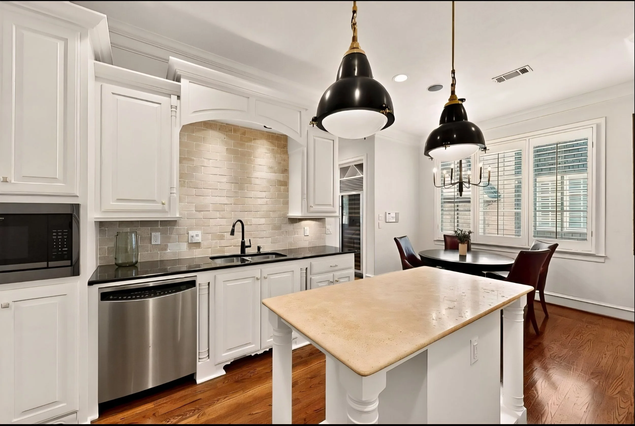 Modern kitchen with white cabinetry, black countertops, and a beige brick backsplash. Contains a black sink, a microwave, a stainless steel dishwasher, pendant lights, and a small dining area with a round table and chairs near windows.