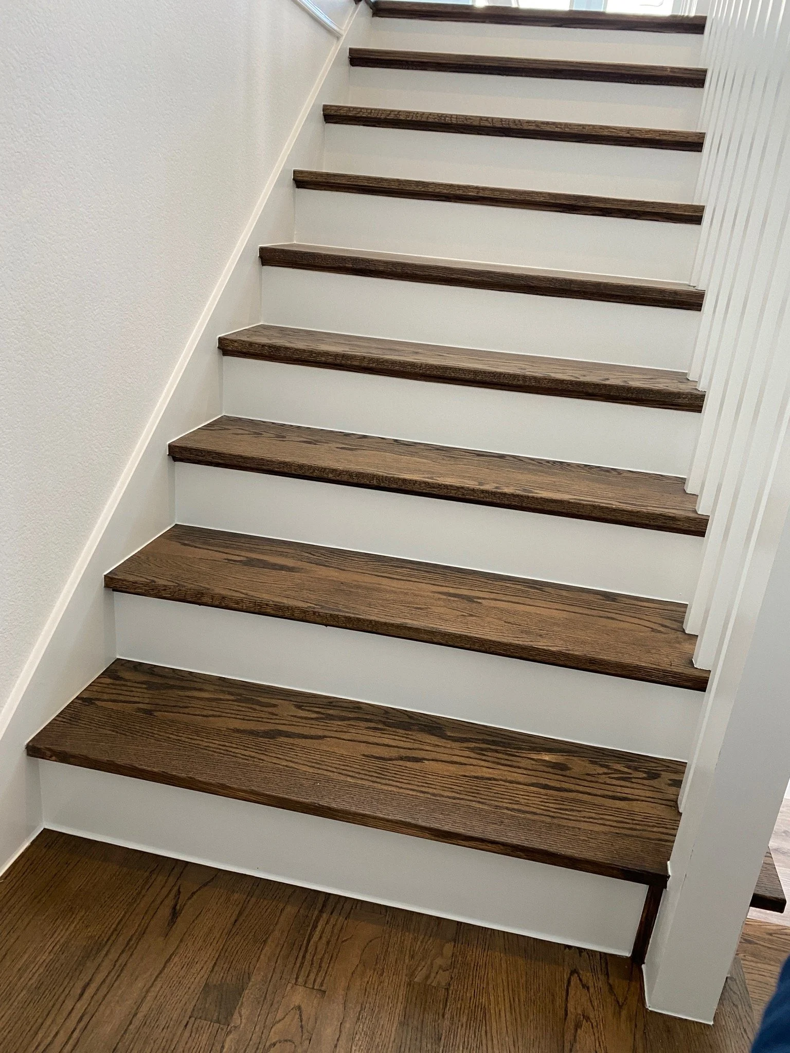 Wooden staircase with dark brown treads and white risers, next to white wall and vertical white paneling.