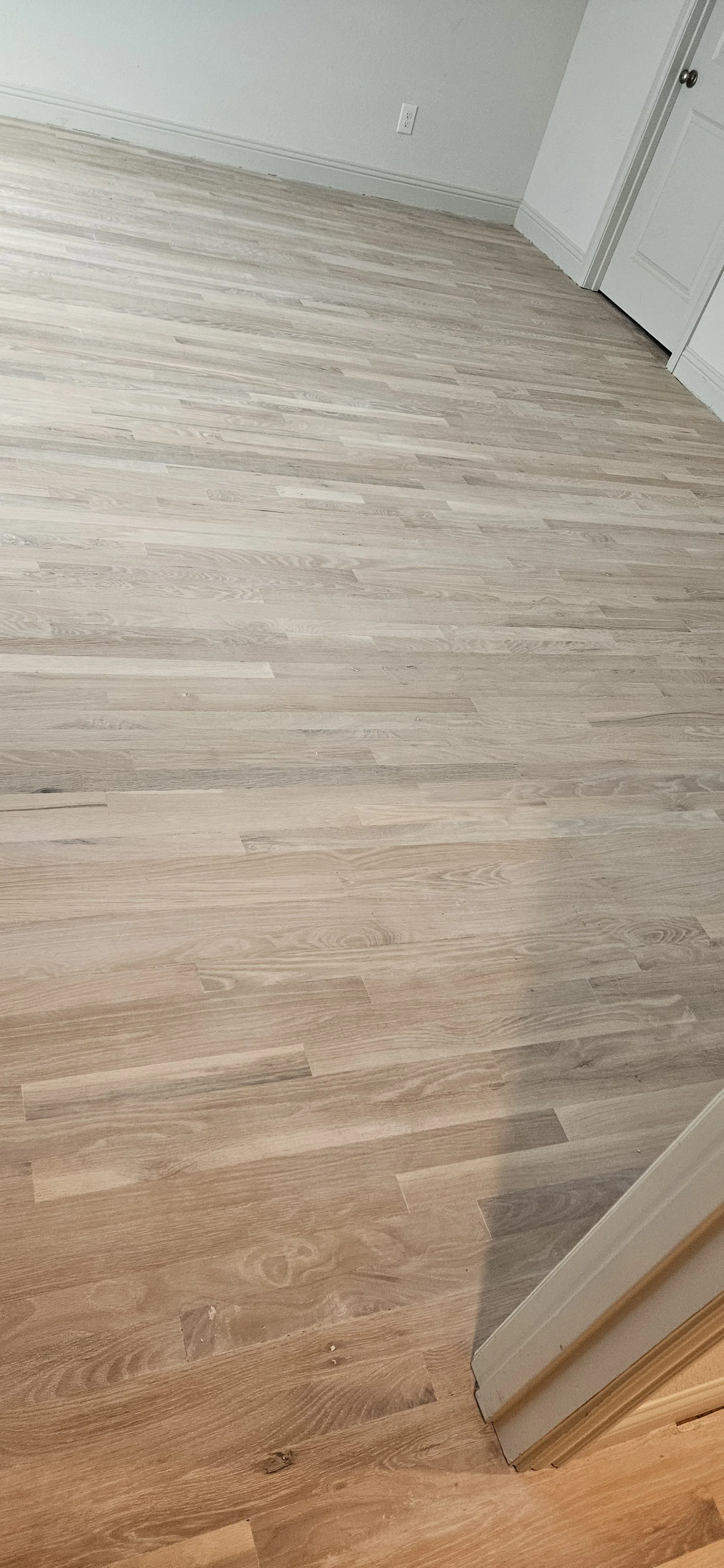Image of a room with light wood laminate flooring, a white wall, electrical outlet, and a door on the right side.