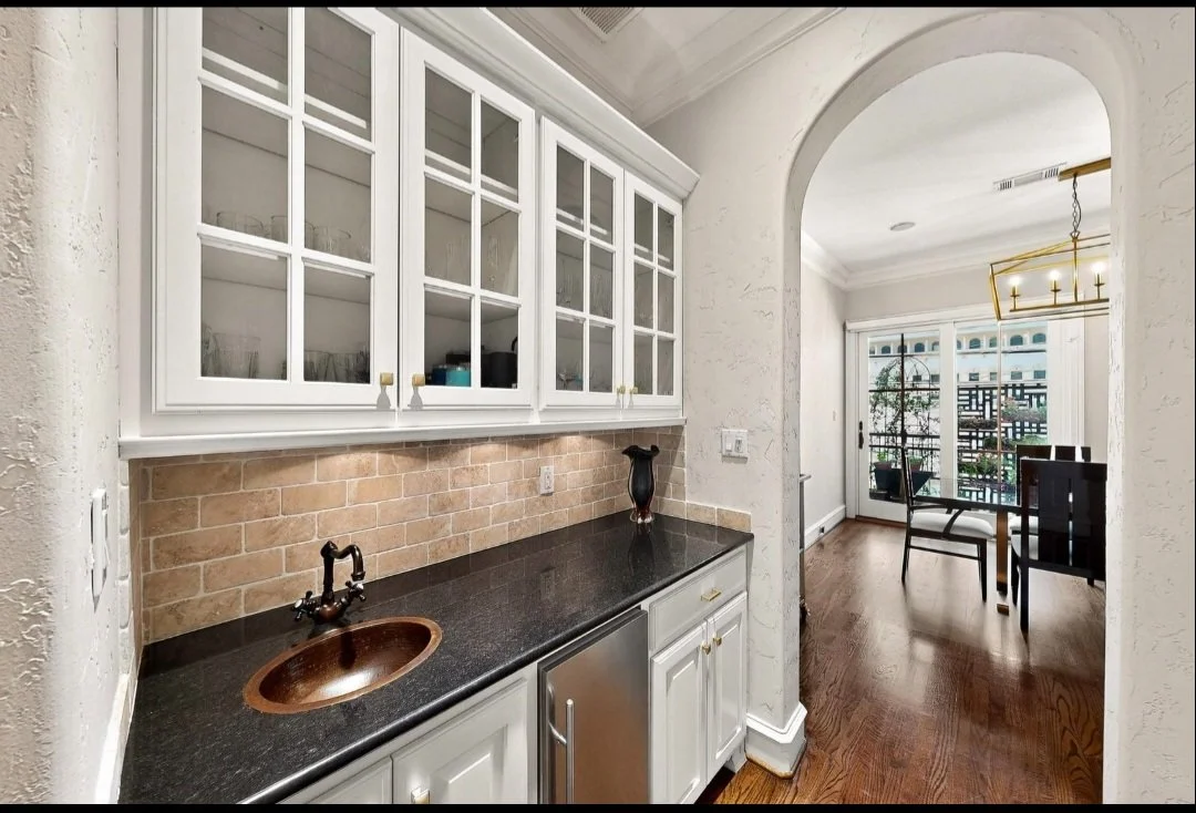 Kitchen wet bar area with white cabinets, dark countertop, brick backsplash, small copper sink, decorative vase, and a view into a dining room with hardwood floor, table and chairs, and a balcony outside.