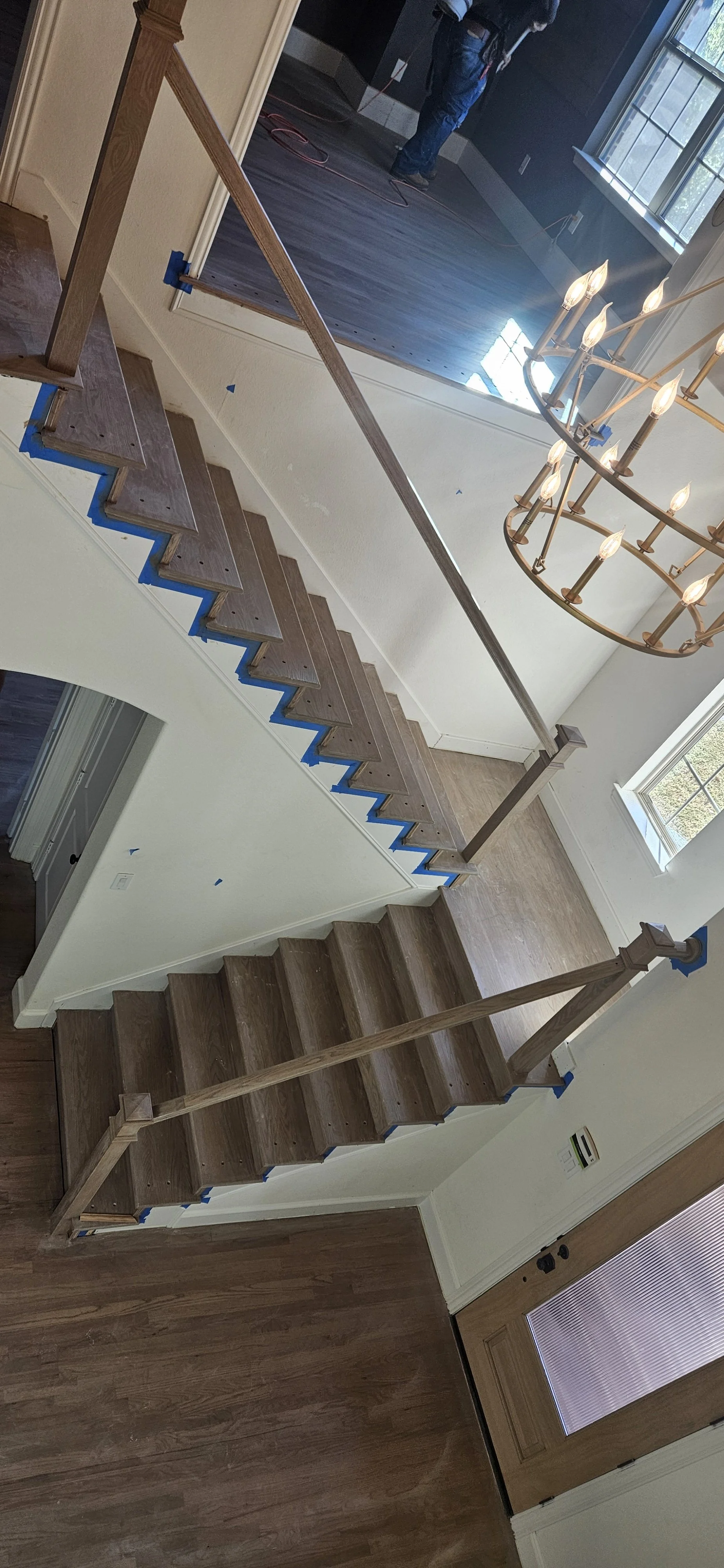Partial view of a wooden staircase under construction with blue painter's tape on the edges, a chandelier with lit bulbs hanging above, and a person at the top working in a room with dark wooden flooring and large windows.