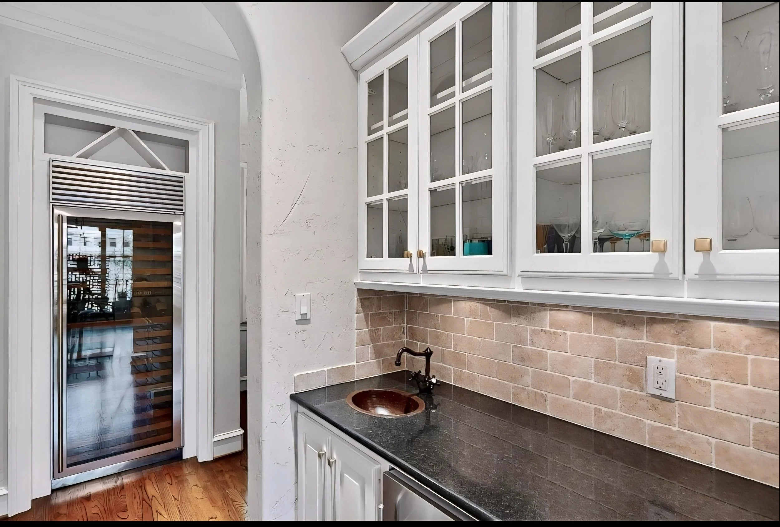 Kitchen with white cabinets, glass-front upper cabinets, black countertop, brick backsplash, small copper sink, and a wine cooler door.