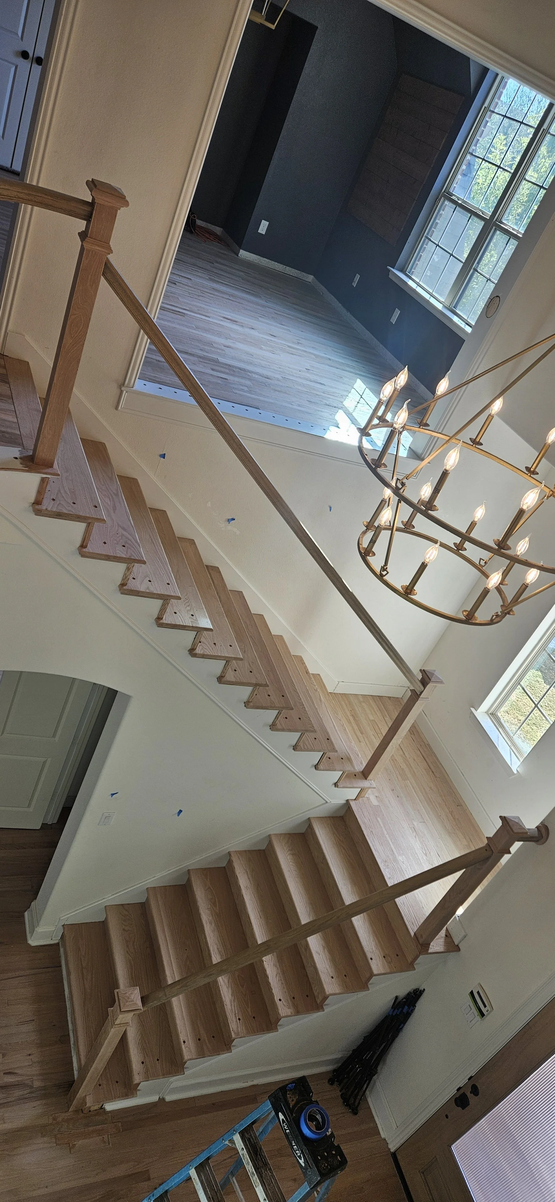 Wooden staircase leading up to a second floor with a chandelier hanging above, and a room in the background with dark walls, large windows, and hardwood flooring.