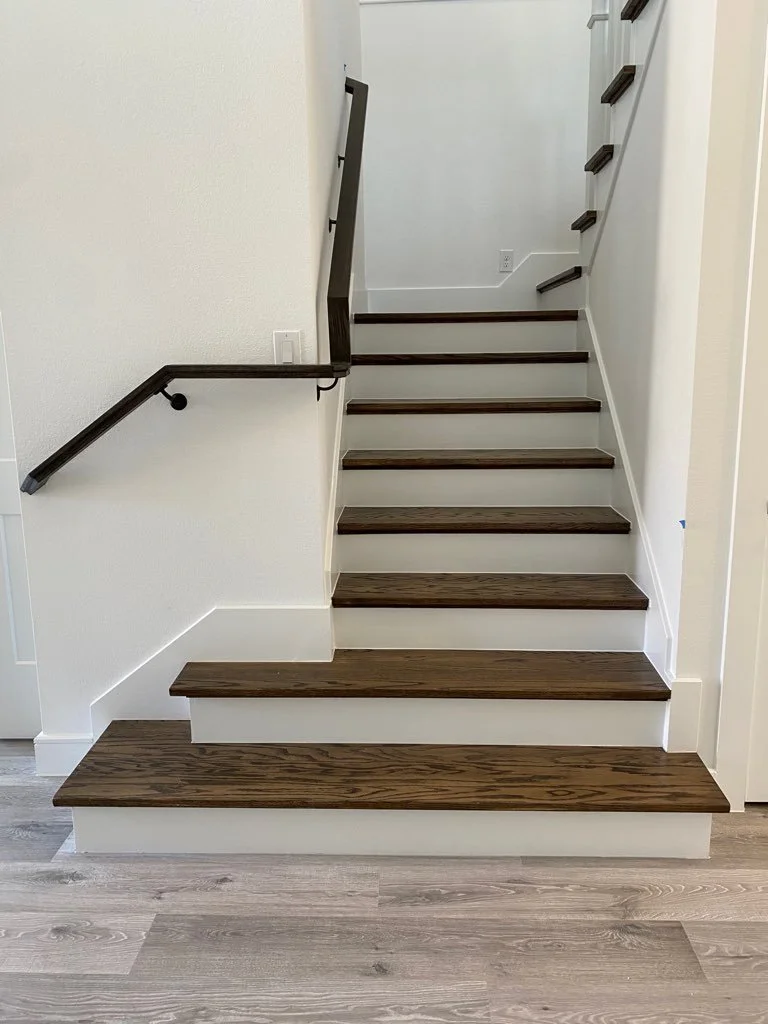 A staircase with dark wood steps and white risers, featuring a dark wood handrail on the left side and small dark wood supports along the right side, against white walls and light-colored flooring.