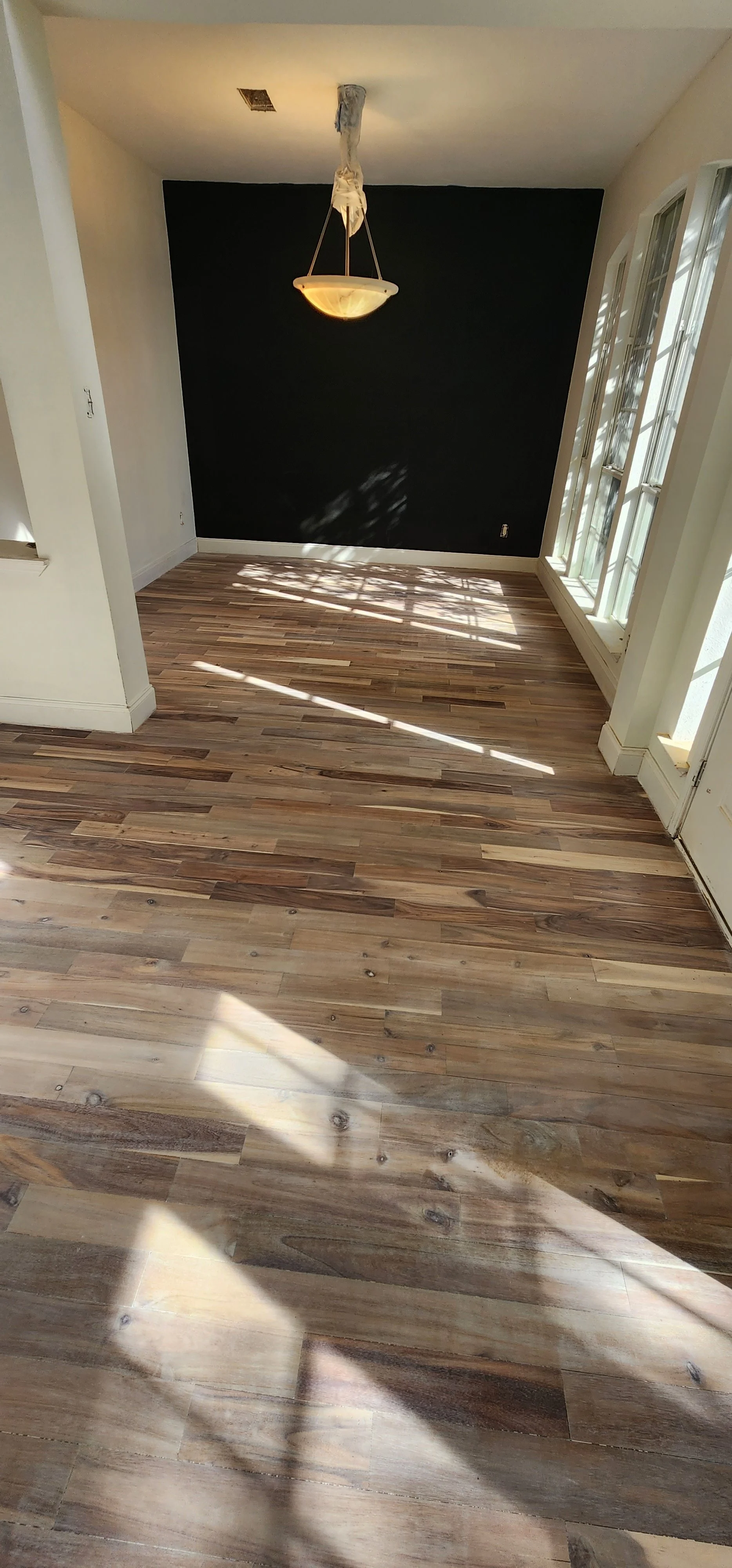 Empty room with hardwood flooring, a black accent wall, a chandelier hanging from the ceiling, and large windows allowing sunlight to cast shadows on the floor.