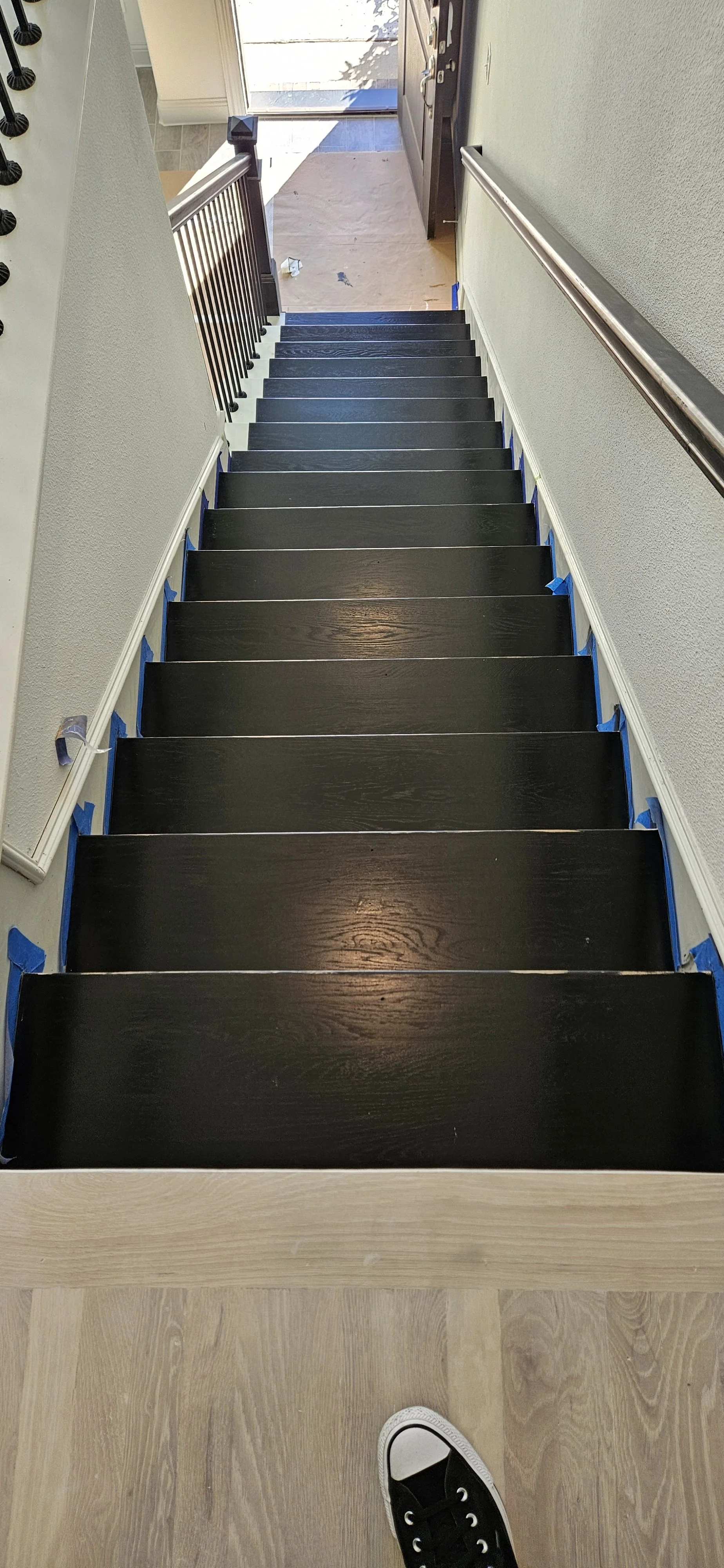 View looking down a staircase with black painted wooden steps, blue painter's tape at the edges, and a person's foot in a black sneaker at the top. The staircase leads to an entrance door with a small front porch area visible outside.