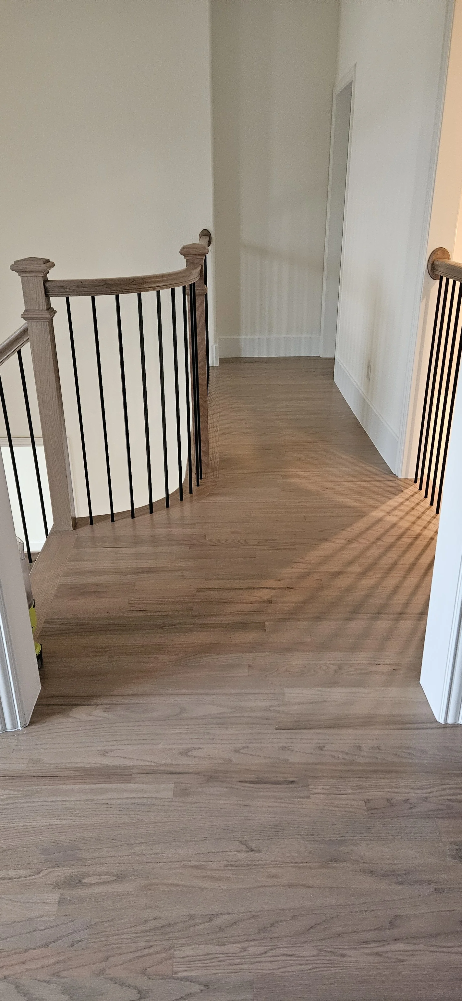 View of a wooden-floored hallway with a staircase railing on the left and a door at the end of the hallway.