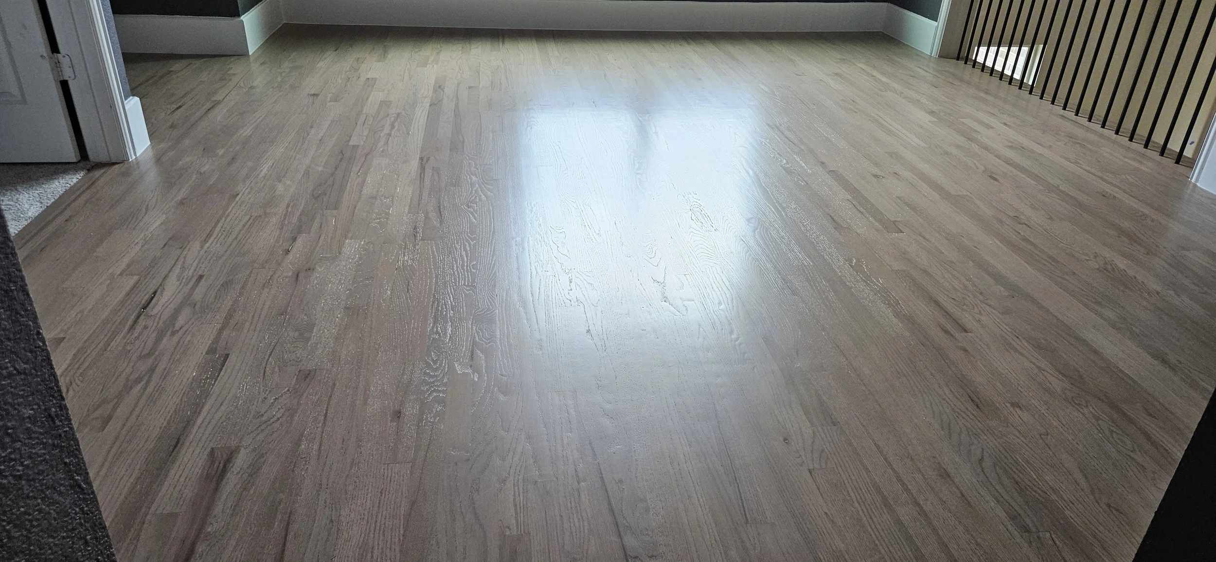 Room with wooden floor, staircase railing on right, doorway on left, and natural light coming through window.