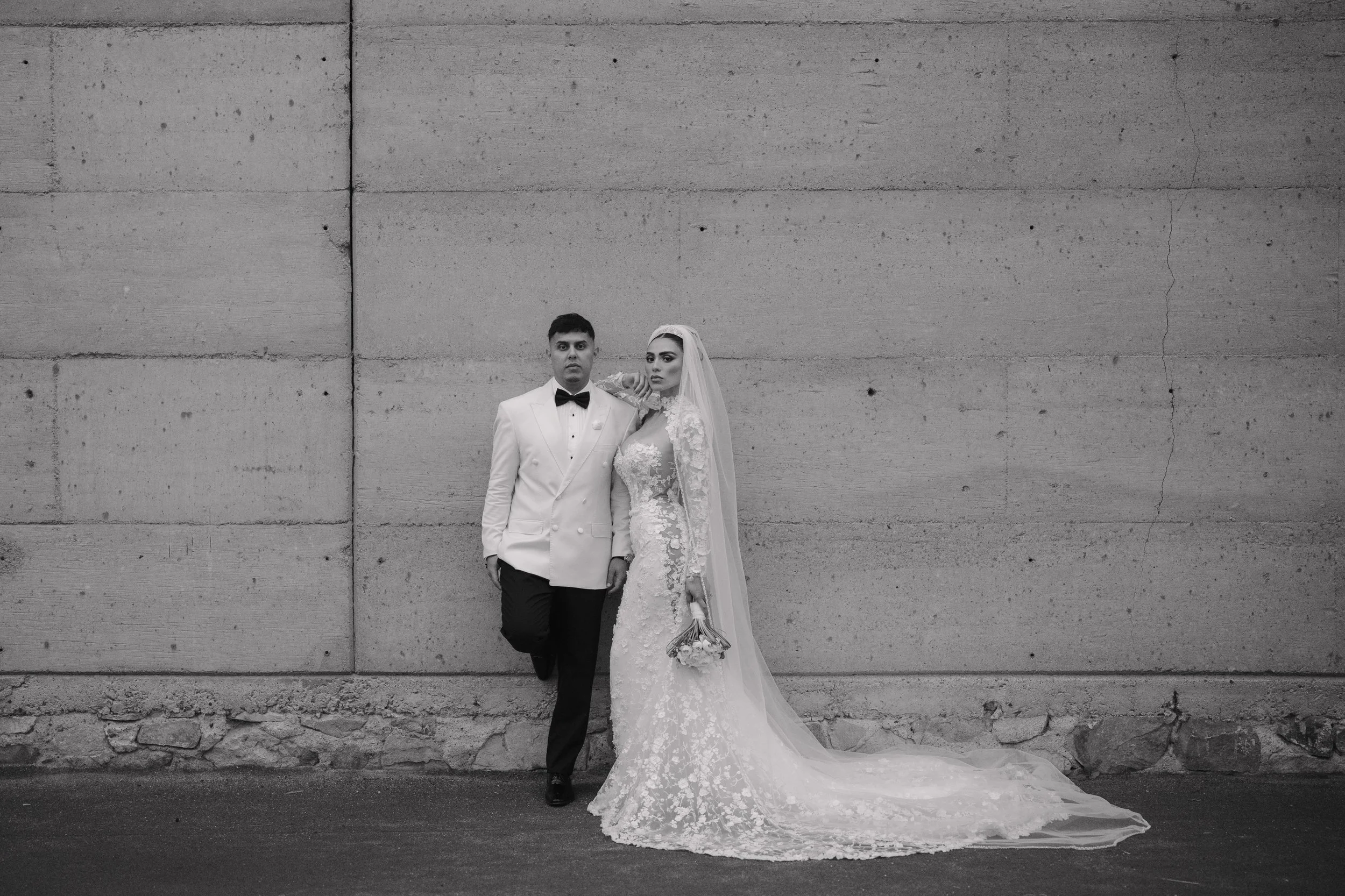 A bride in a lace wedding gown and veil stands next to a groom in a tuxedo with a bow tie, against a large concrete wall. Wedding Photography in Valle de Guadalupe, Ensenada, Baja California.