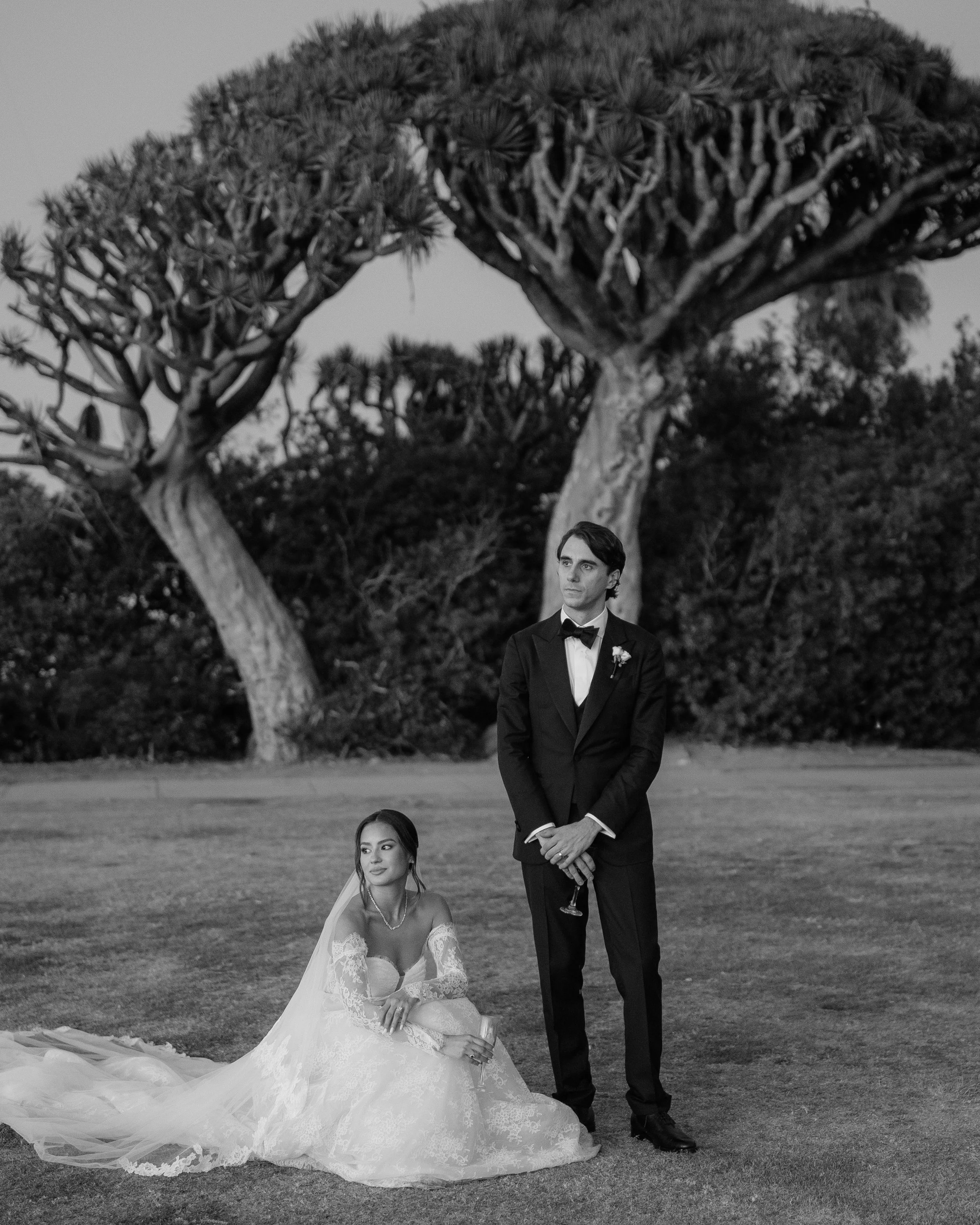 A black and white photo of a bride sitting on the grass wearing a wedding dress with a long train, and a groom standing beside her in a tuxedo, holding a drink, outdoors with trees in the background. Wedding Photography in Valle de Guadalupe.