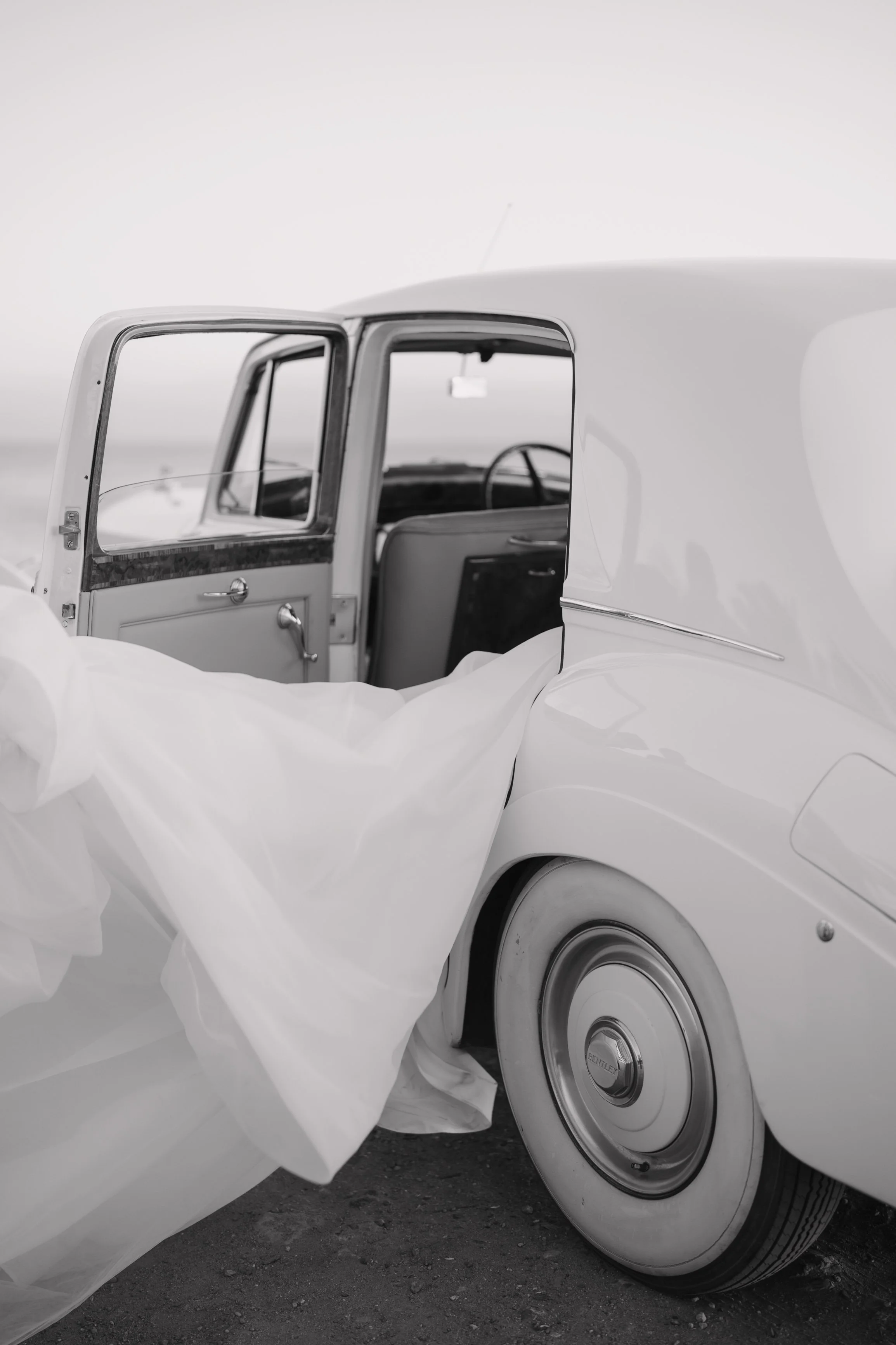 A vintage white car with an open door and a white fabric draped over it, captured in black and white, set against a plain background. Wedding Photography in Valle de Guadalupe, Ensenada, Baja California.