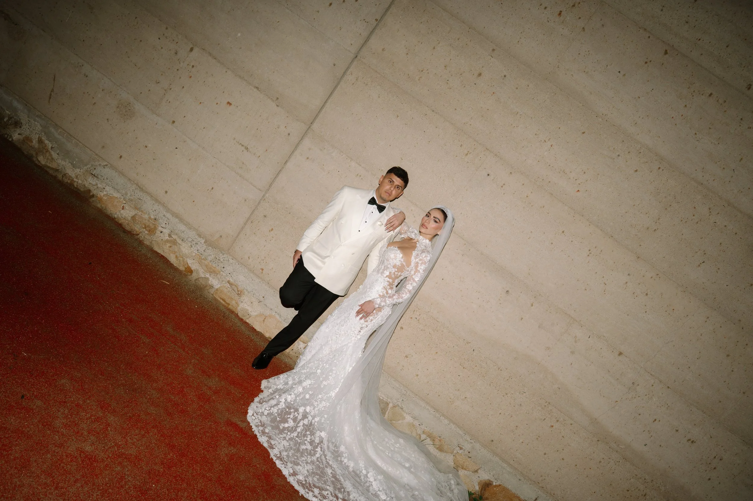 A bride and groom dressed in wedding attire standing against a concrete wall with a red carpeted floor. Wedding Photography in Valle de Guadalupe, Ensenada, Baja California.