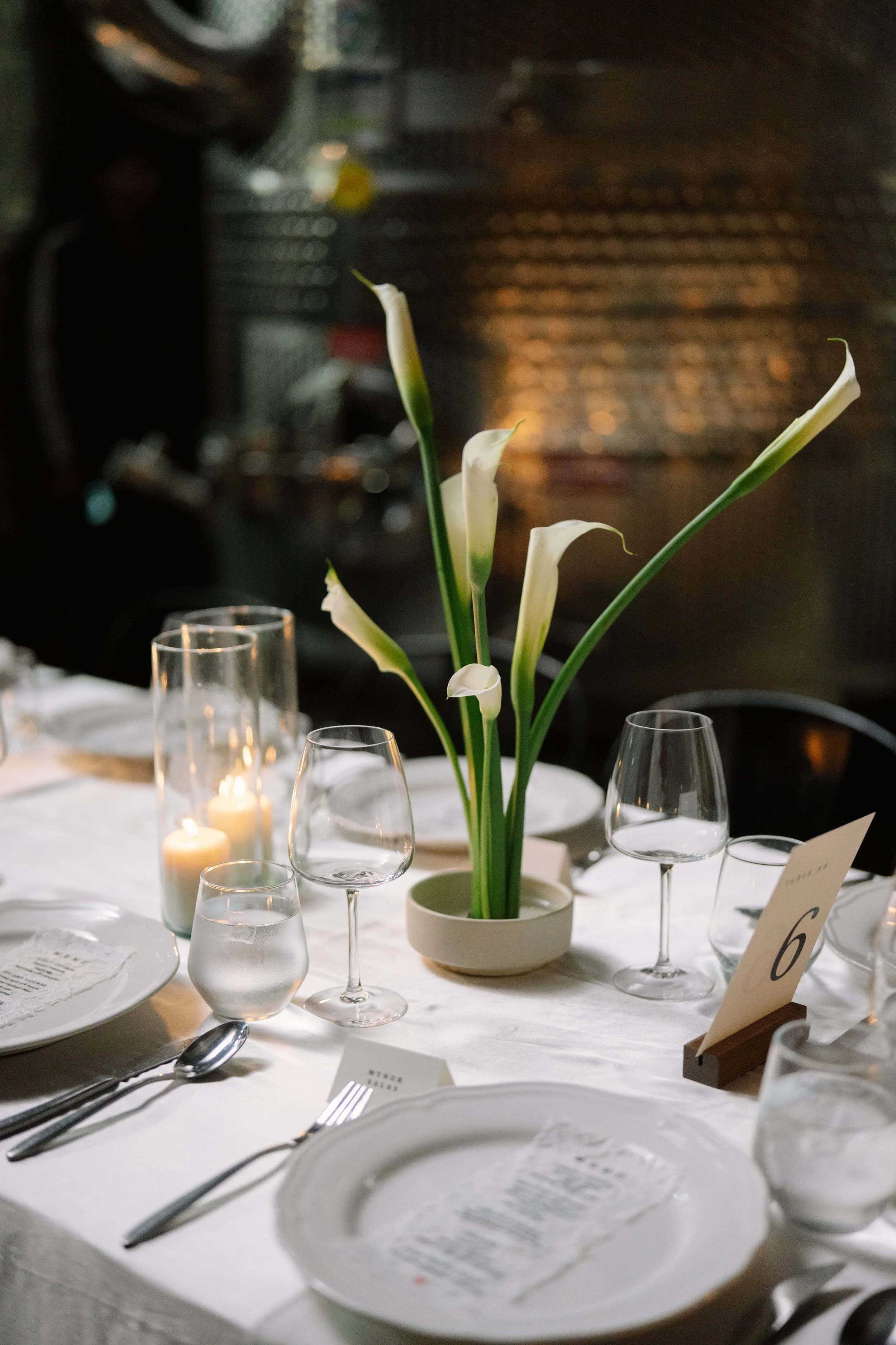 Elegant dining table with white tablecloth, floral centerpiece of white calla lilies, candles, glassware, and table number '6' in a dimly lit setting. Wedding Photography in Valle de Guadalupe, Ensenada, Baja California.