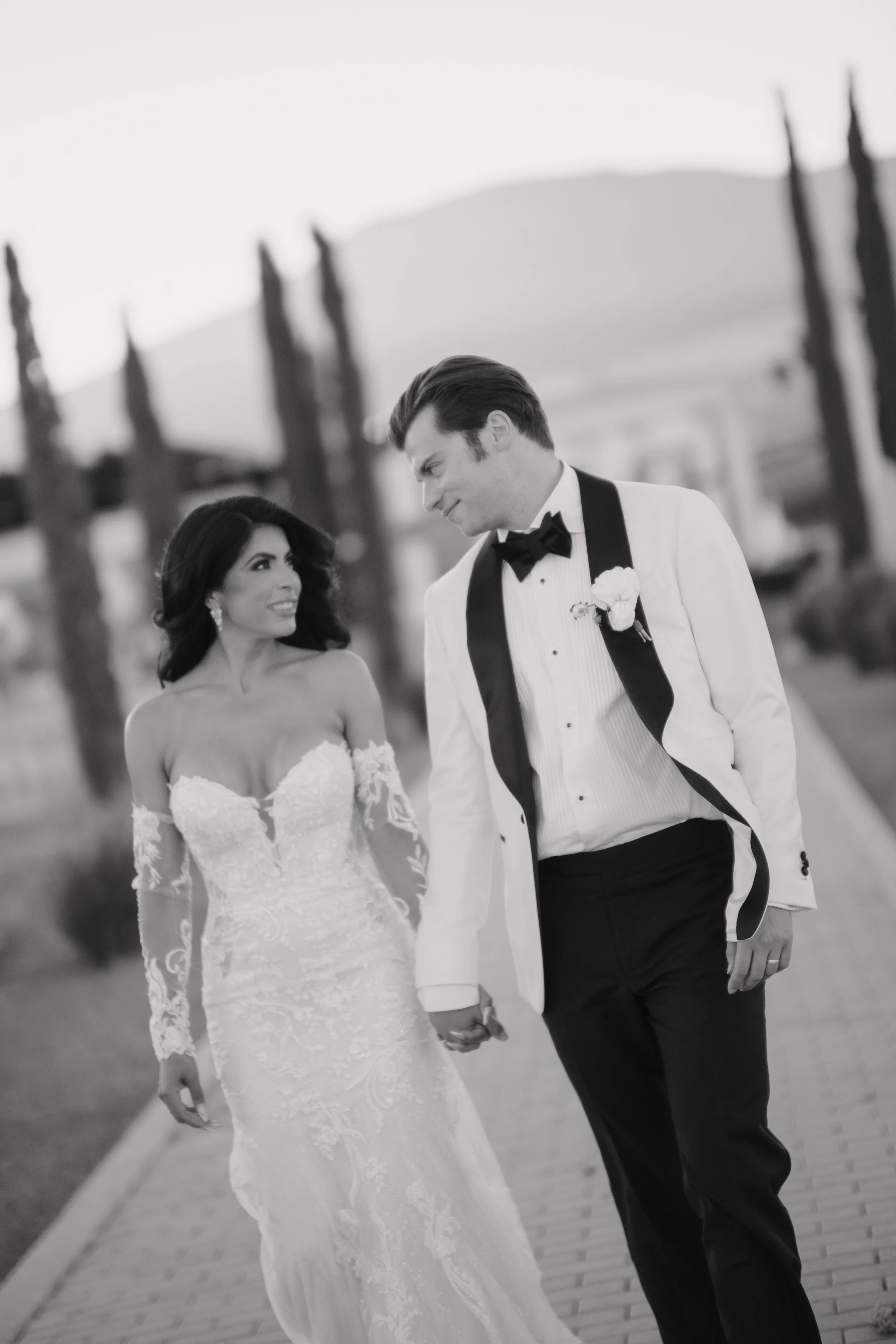 A black-and-white photo of a bride and groom walking hand-in-hand outdoors, with trees and a mountain in the background. Wedding Photography in Valle de Guadalupe, Ensenada, Baja California.