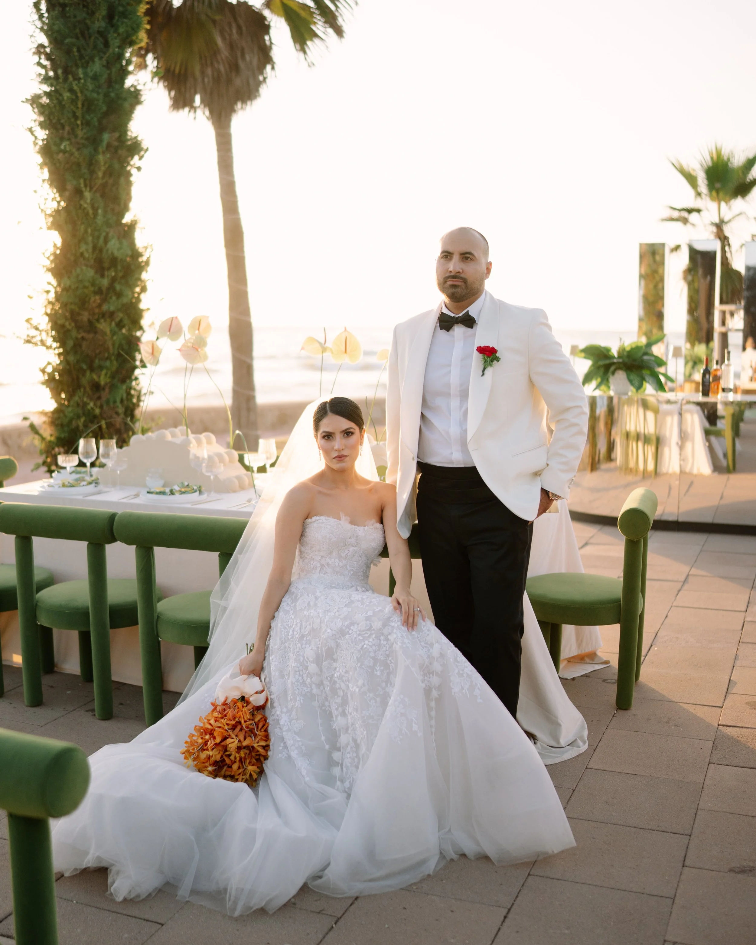 A bride in a white wedding gown sitting with a bouquet of orange flowers, and a groom in a white tuxedo with a black bow tie and red boutonniere, standing beside her on a beachside outdoor wedding setup during sunset. Wedding Photography in Ensenada.
