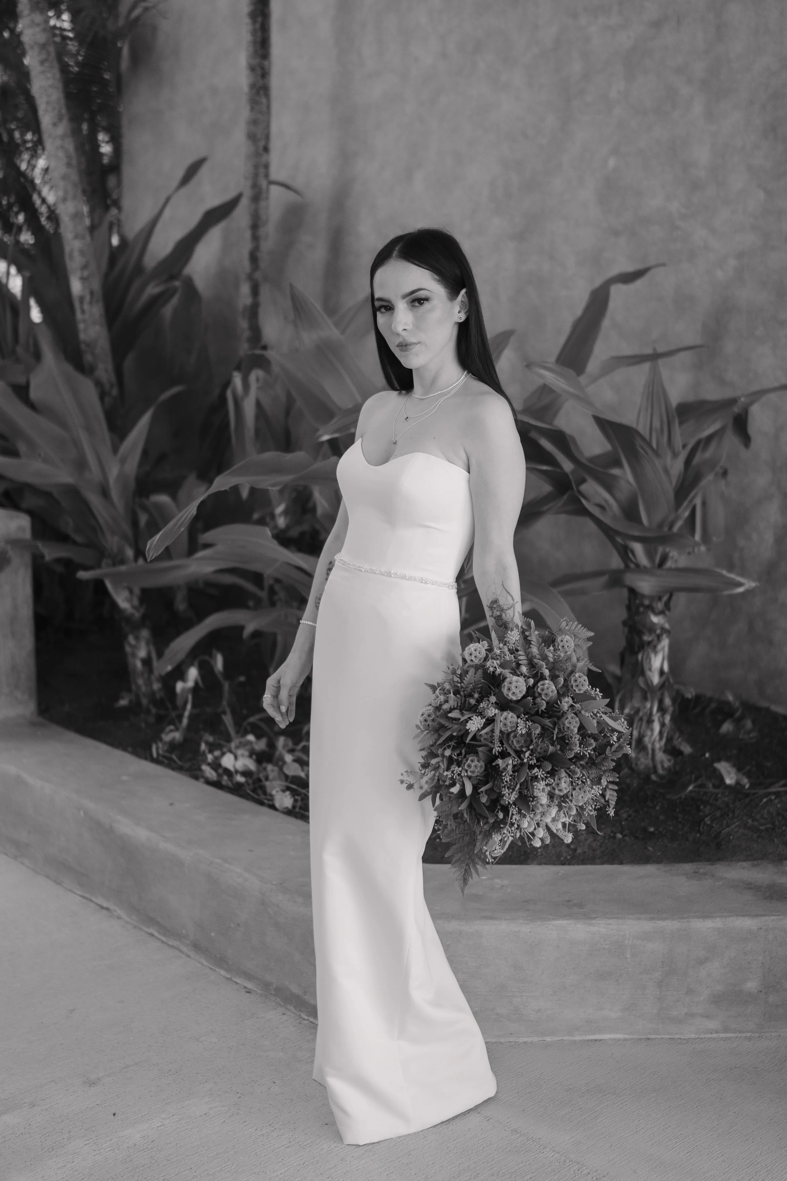 A woman in a white wedding dress holding a bouquet of flowers standing outdoors near large leafy plants. Wedding Photography in Valle de Guadalupe, Ensenada, Baja California.