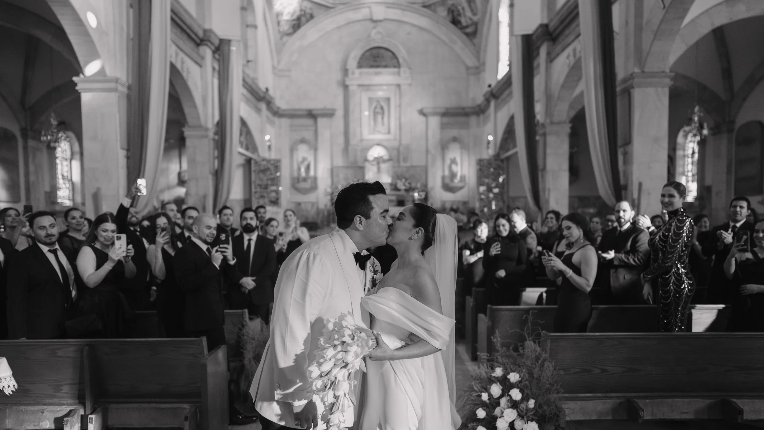 A bride and groom kissing during their wedding ceremony inside a church, with guests taking photos and watching. Wedding Photography in Valle de Guadalupe, Ensenada, Baja California.