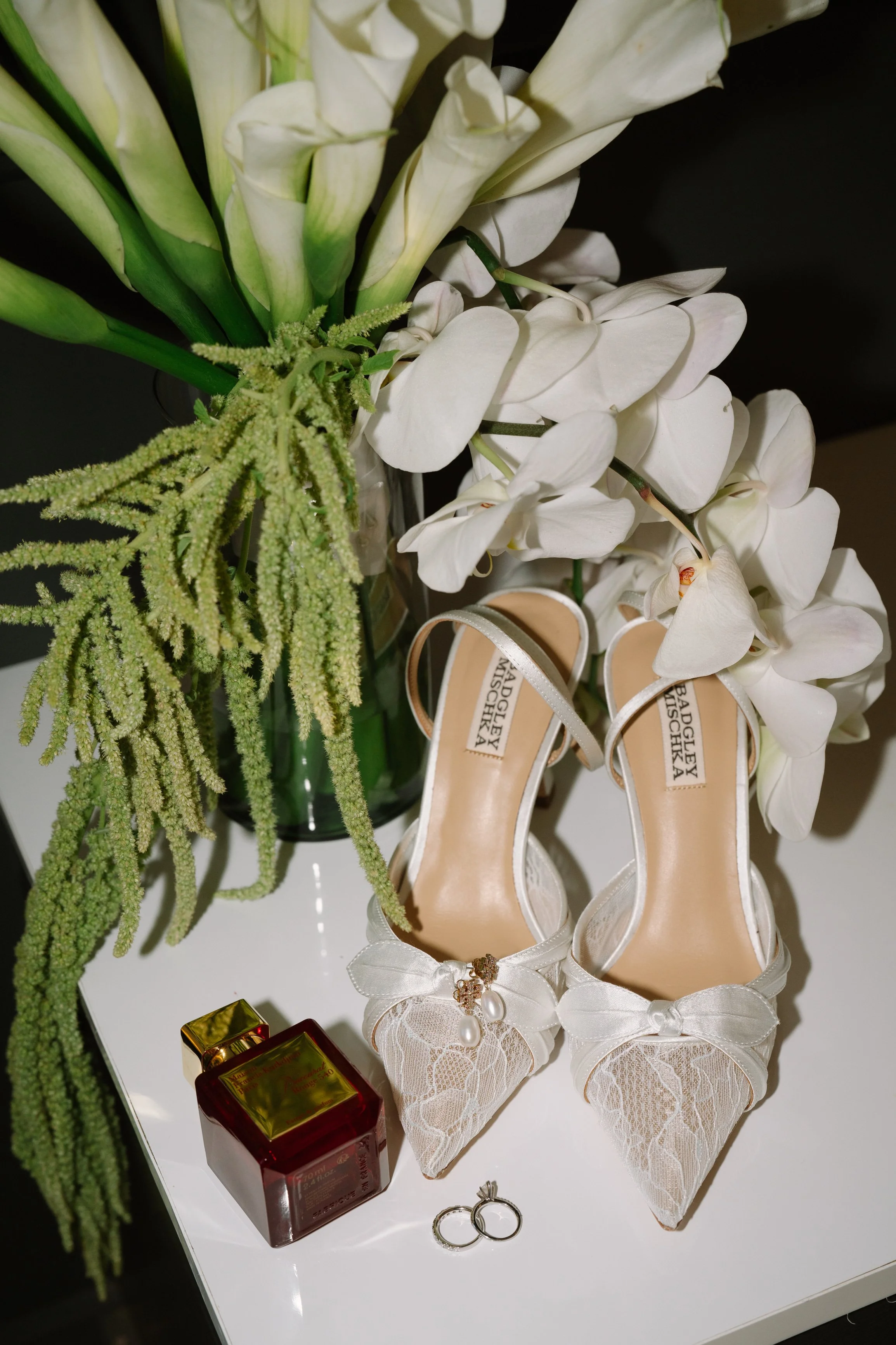 White wedding shoes with lace and bows, placed beside wedding rings, next to a bottle of perfume, all on a white table with a bouquet of white flowers and greenery. Wedding Photography in Valle de Guadalupe, Ensenada, Baja California.