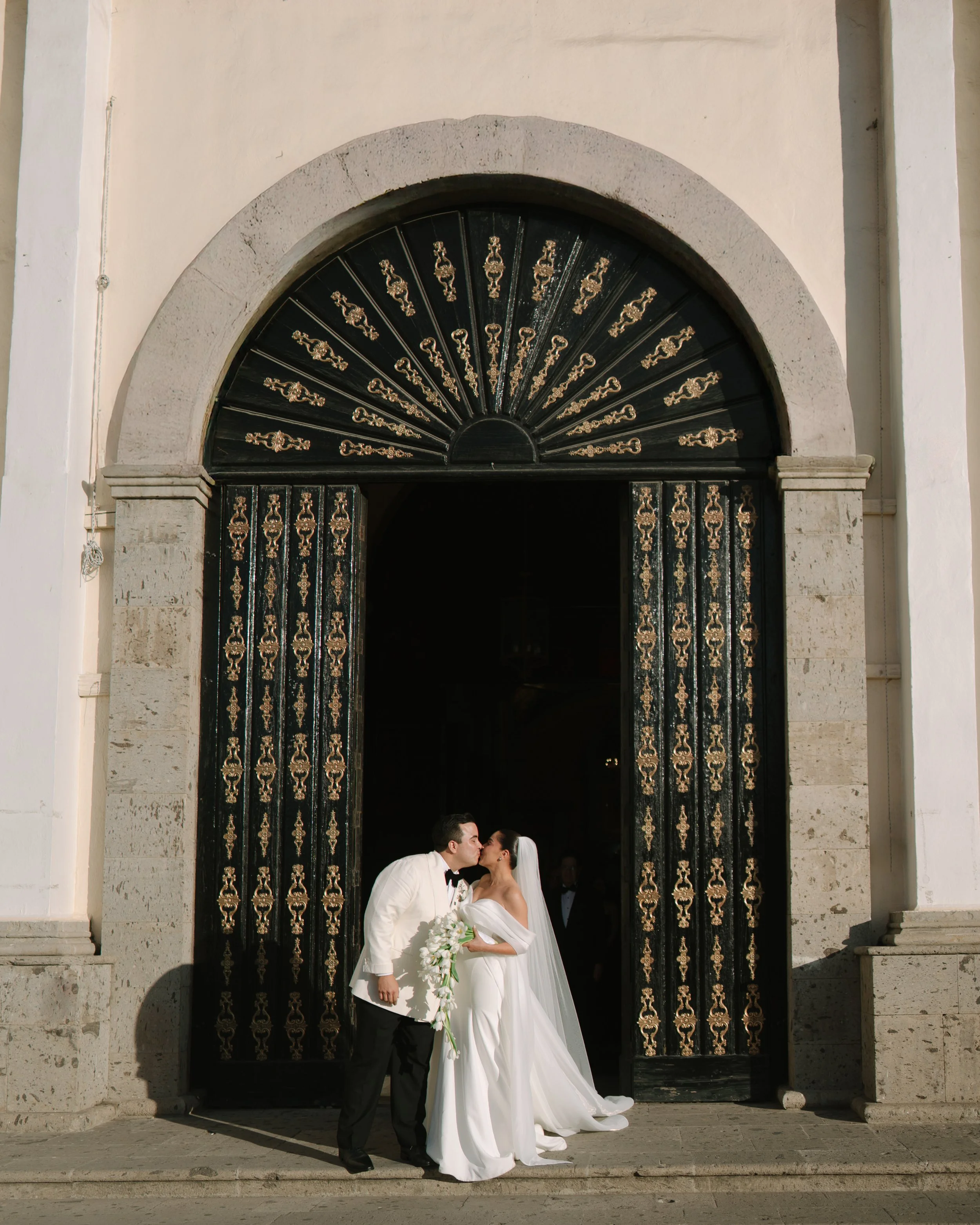 A bride and groom kissing in front of a large ornate black and gold door outside a church. Wedding Photography in Valle de Guadalupe, Ensenada, Baja California.