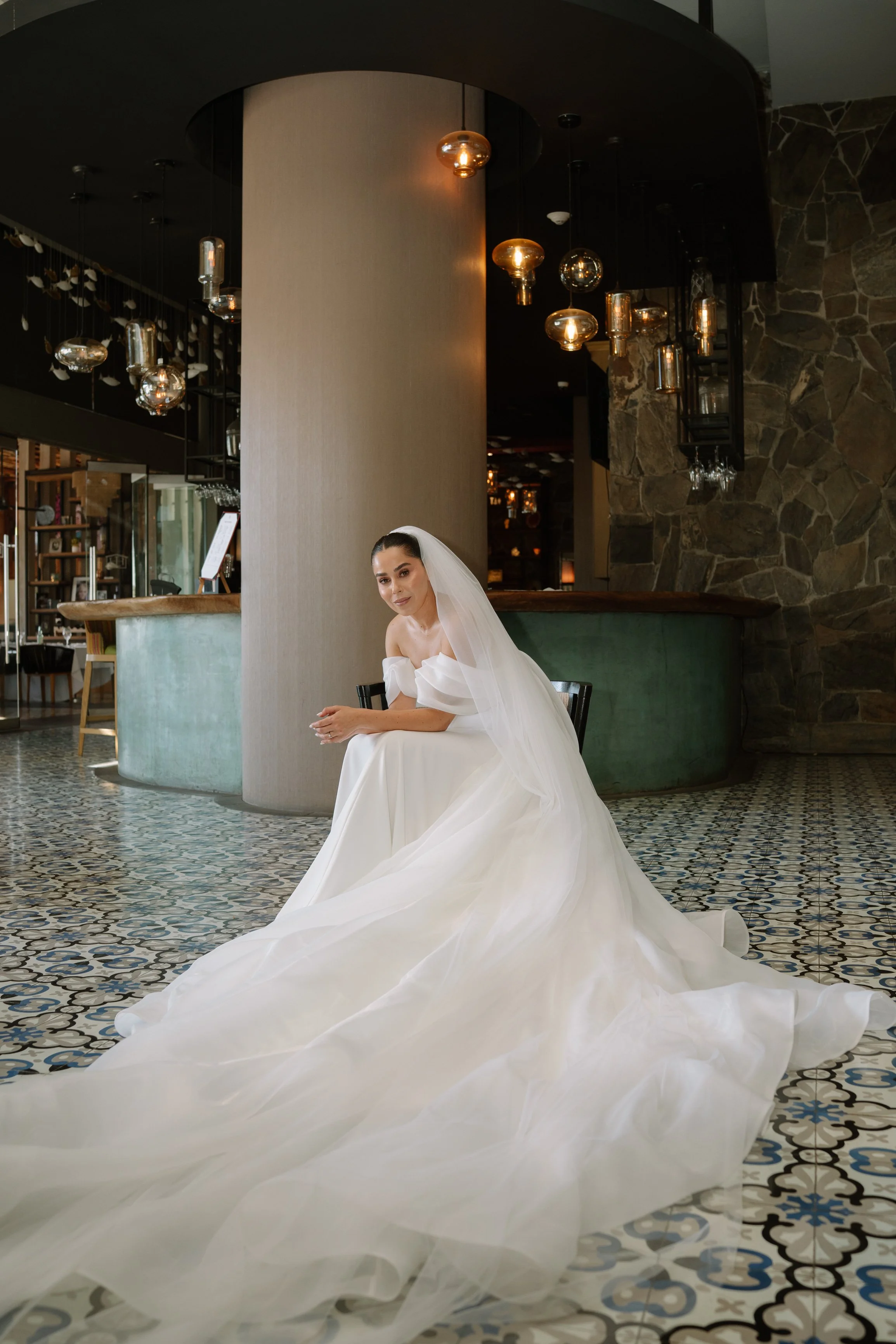 A woman wearing a white wedding gown and veil sitting on a black chair in a modern indoor setting with patterned tile floor and decorative hanging lights. Wedding Photography in Valle de Guadalupe, Ensenada, Baja California.