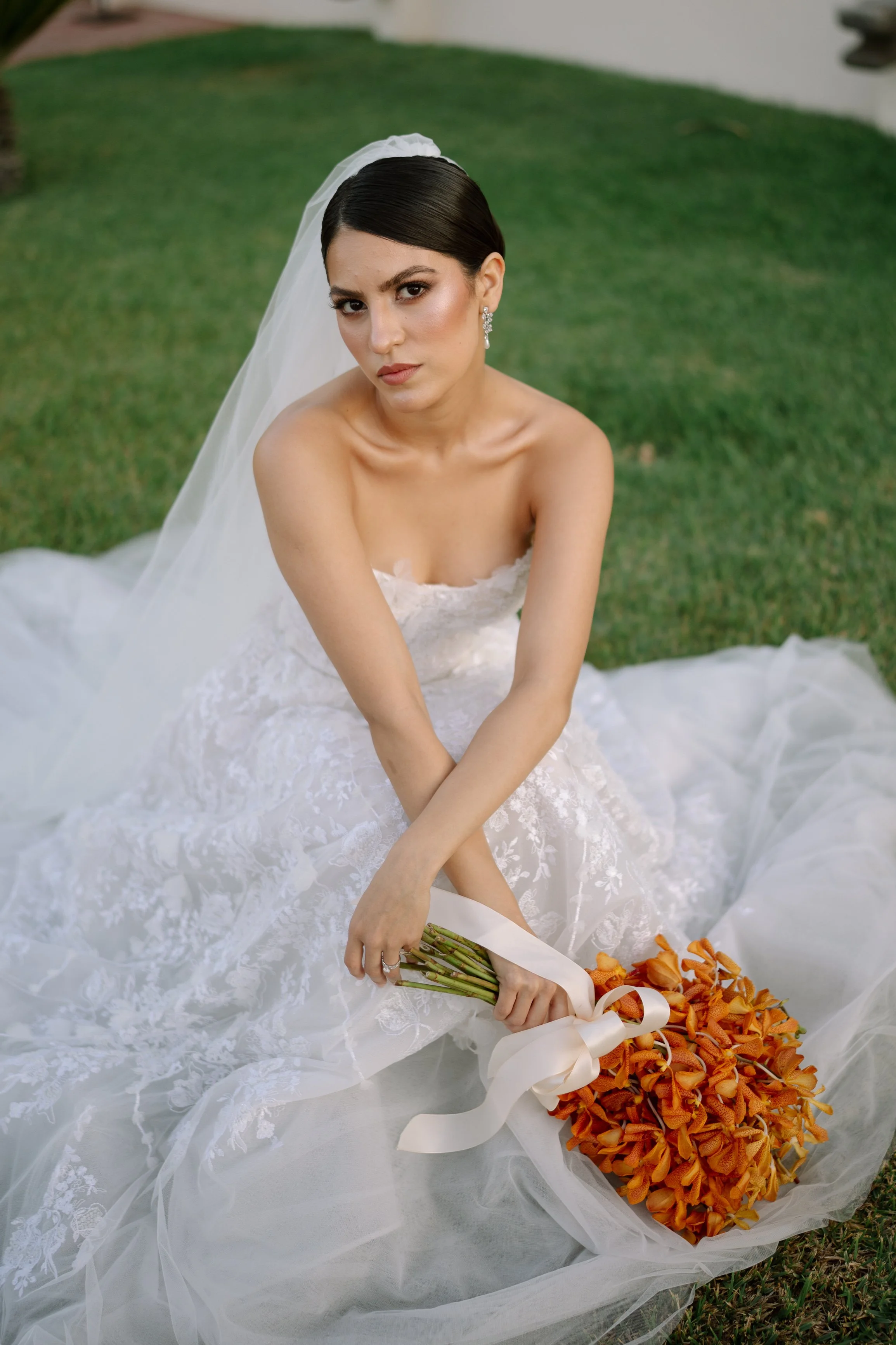 Bride sitting on grass in a wedding dress holding a bouquet of orange flowers with a white ribbon. Wedding Photography in Valle de Guadalupe, Ensenada, Baja California.