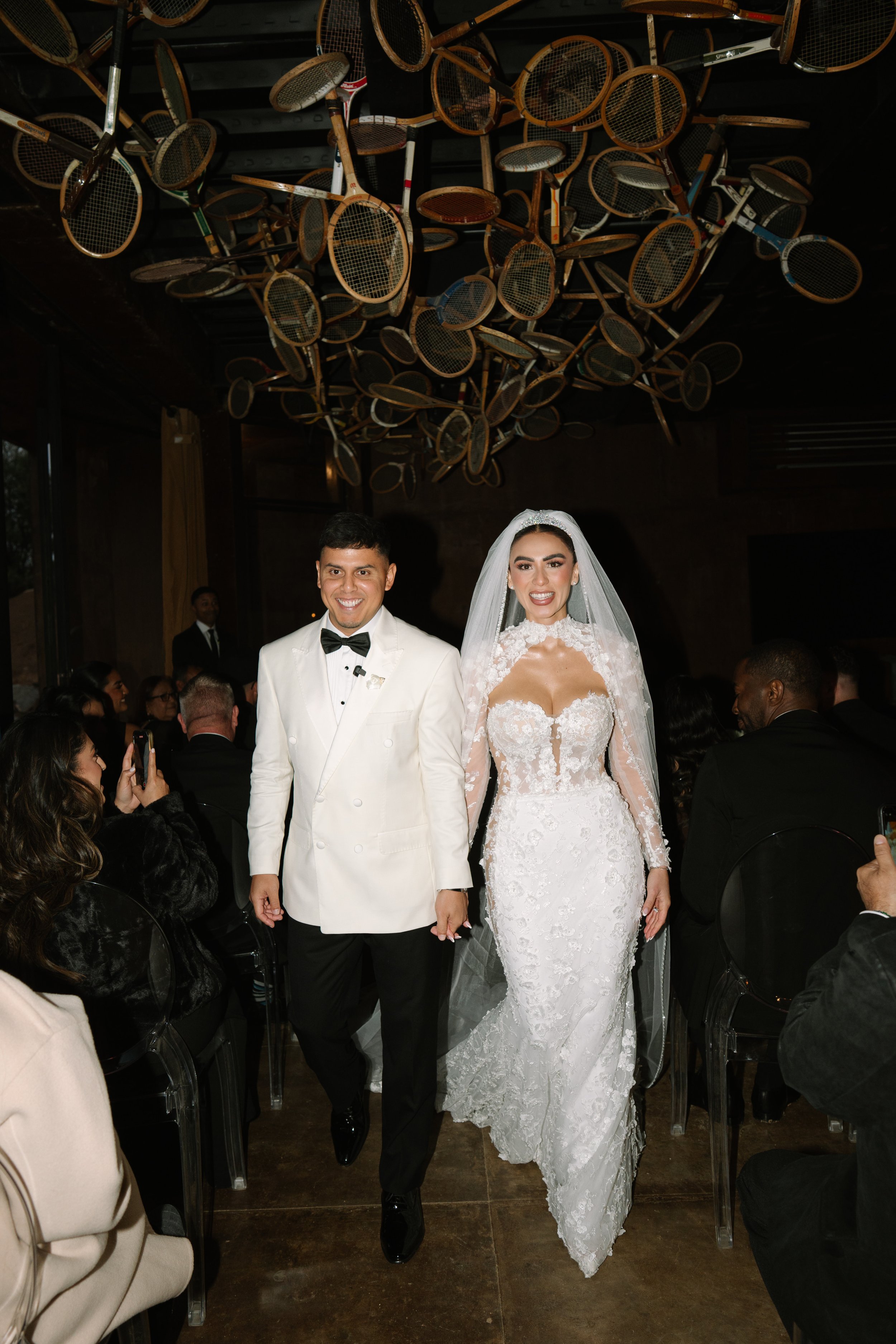 A bride and groom walking down the aisle during their wedding reception, surrounded by seated guests, with a ceiling decorated with multiple tennis rackets. Wedding Photography in Valle de Guadalupe, Ensenada, Baja California.