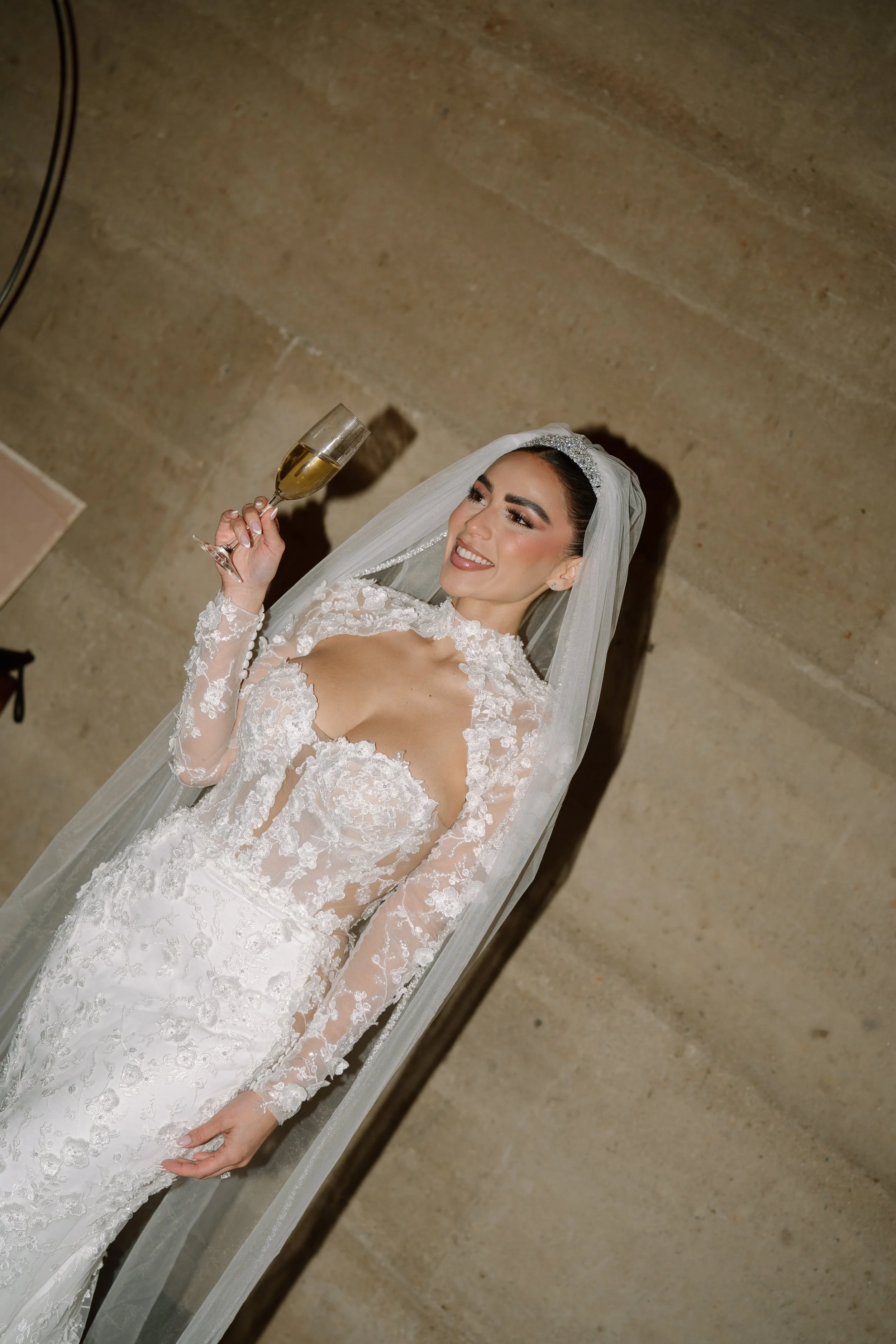 A smiling bride in a lace wedding dress holding a glass of champagne. Wedding Photography in Valle de Guadalupe, Ensenada, Baja California.
