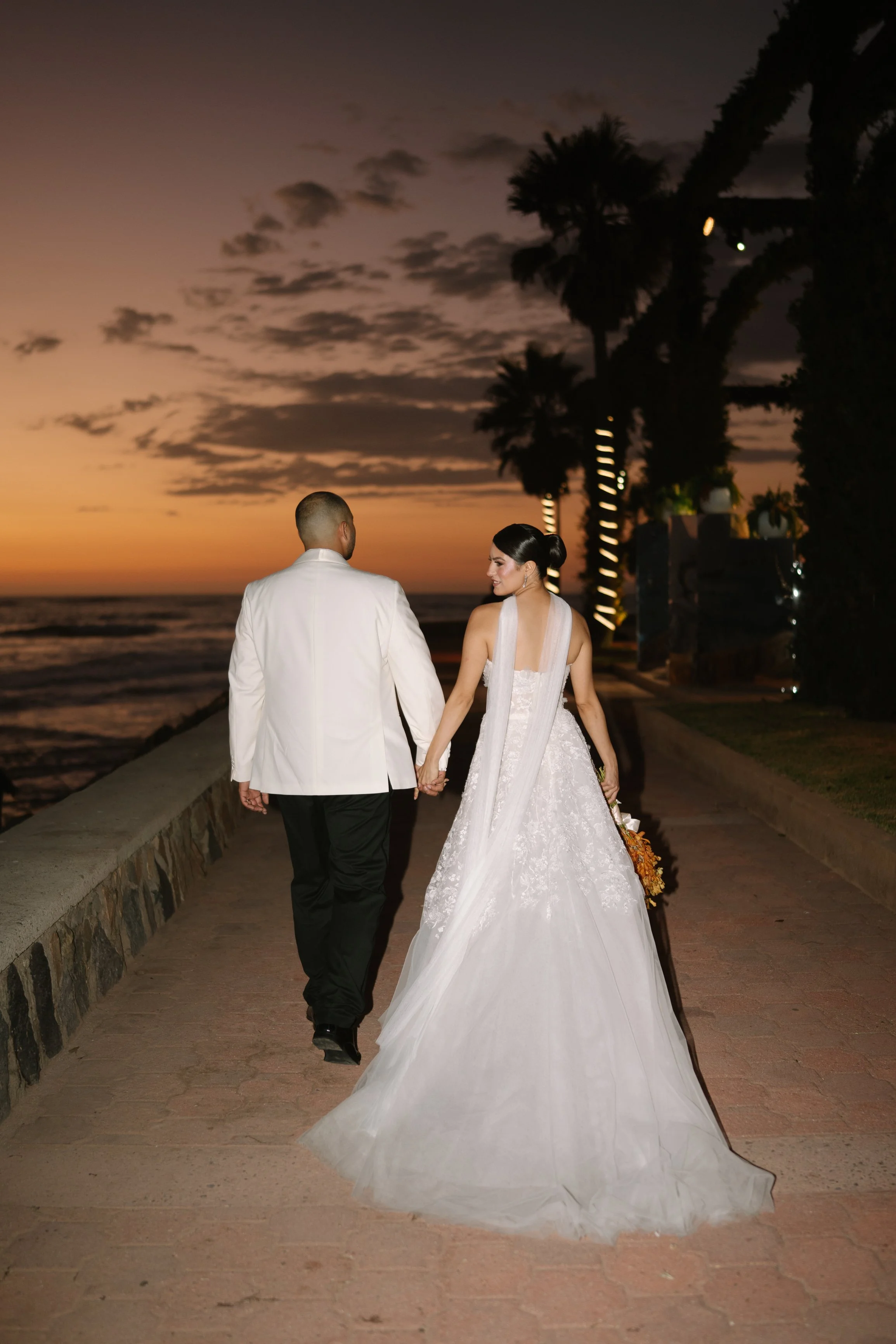 A bride and groom walk hand-in-hand during sunset on a beachside promenade, with palm trees lining the path. Wedding Photography in Valle de Guadalupe, Ensenada, Baja California.