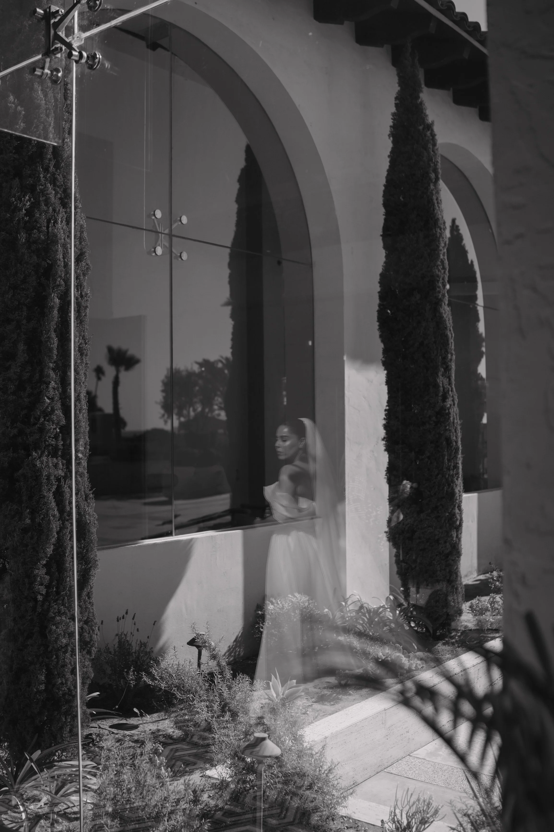 A bride in a white wedding dress and veil looking out through a large window of a building, with tall trees and plants outside, reflected in the glass. Wedding Photography in Valle de Guadalupe, Ensenada, Baja California.