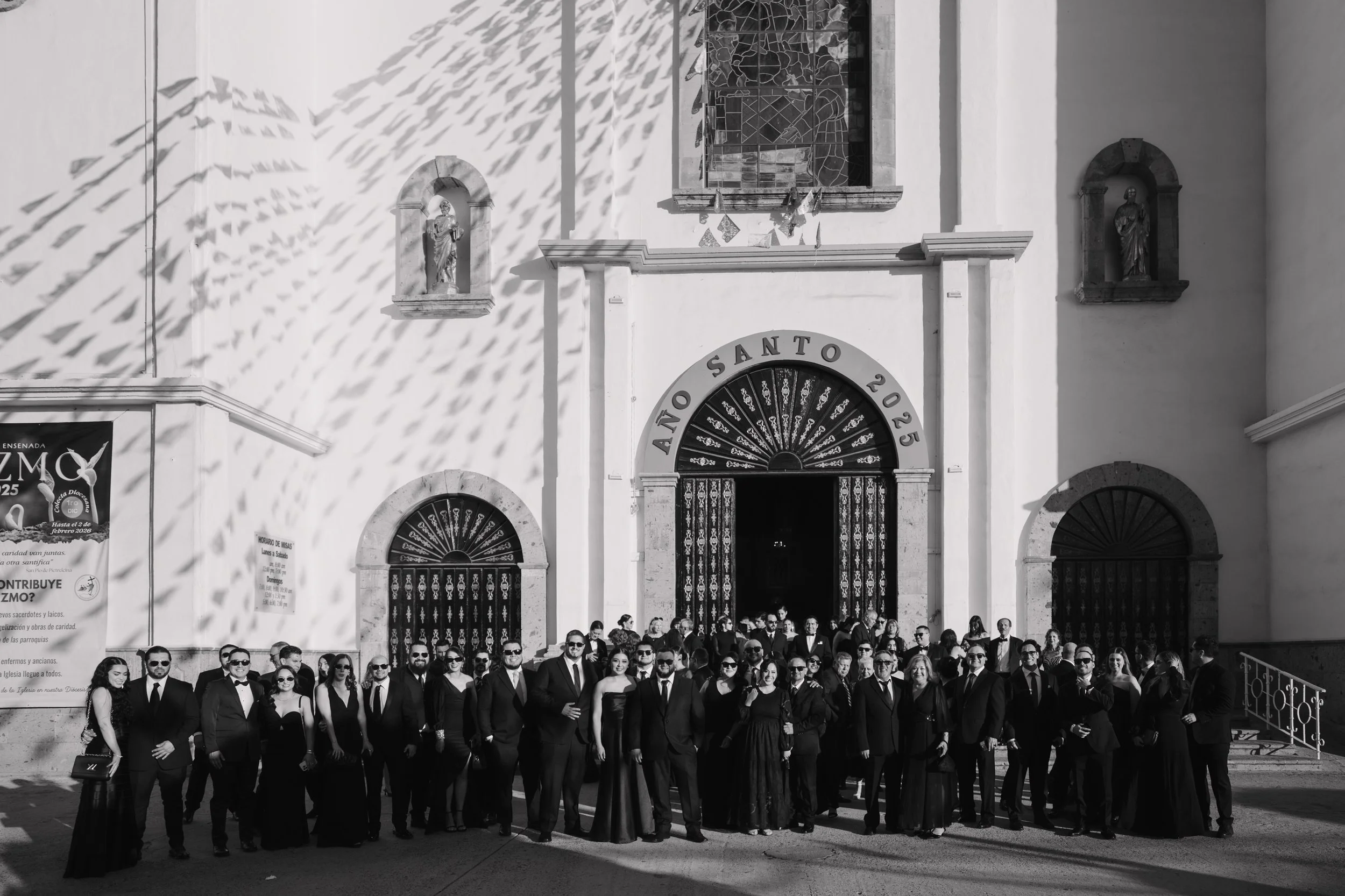 A group of people dressed in formal black attire standing outside a church for a special occasion, with sunlight casting shadows on the building. Wedding Photography in Valle de Guadalupe, Ensenada, Baja California.