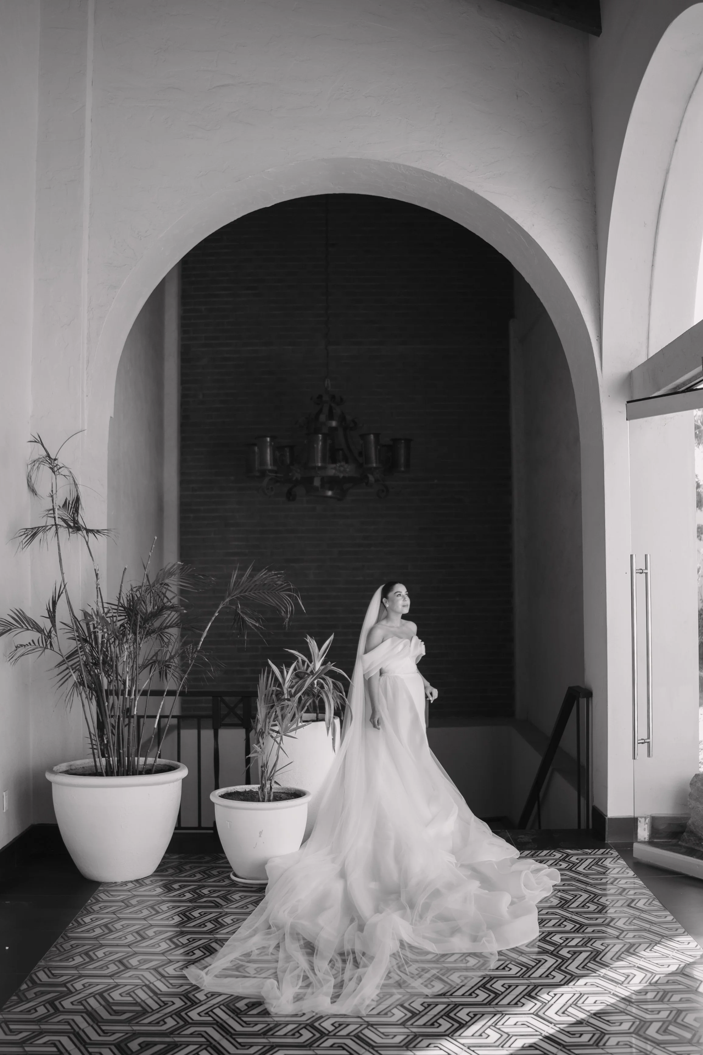 Black and white photo of a bride in a wedding dress standing indoors near large potted plants with a patterned rug underneath, arched doorway, and a chandelier hanging from the ceiling. Wedding Photography in Valle de Guadalupe, Ensenada.