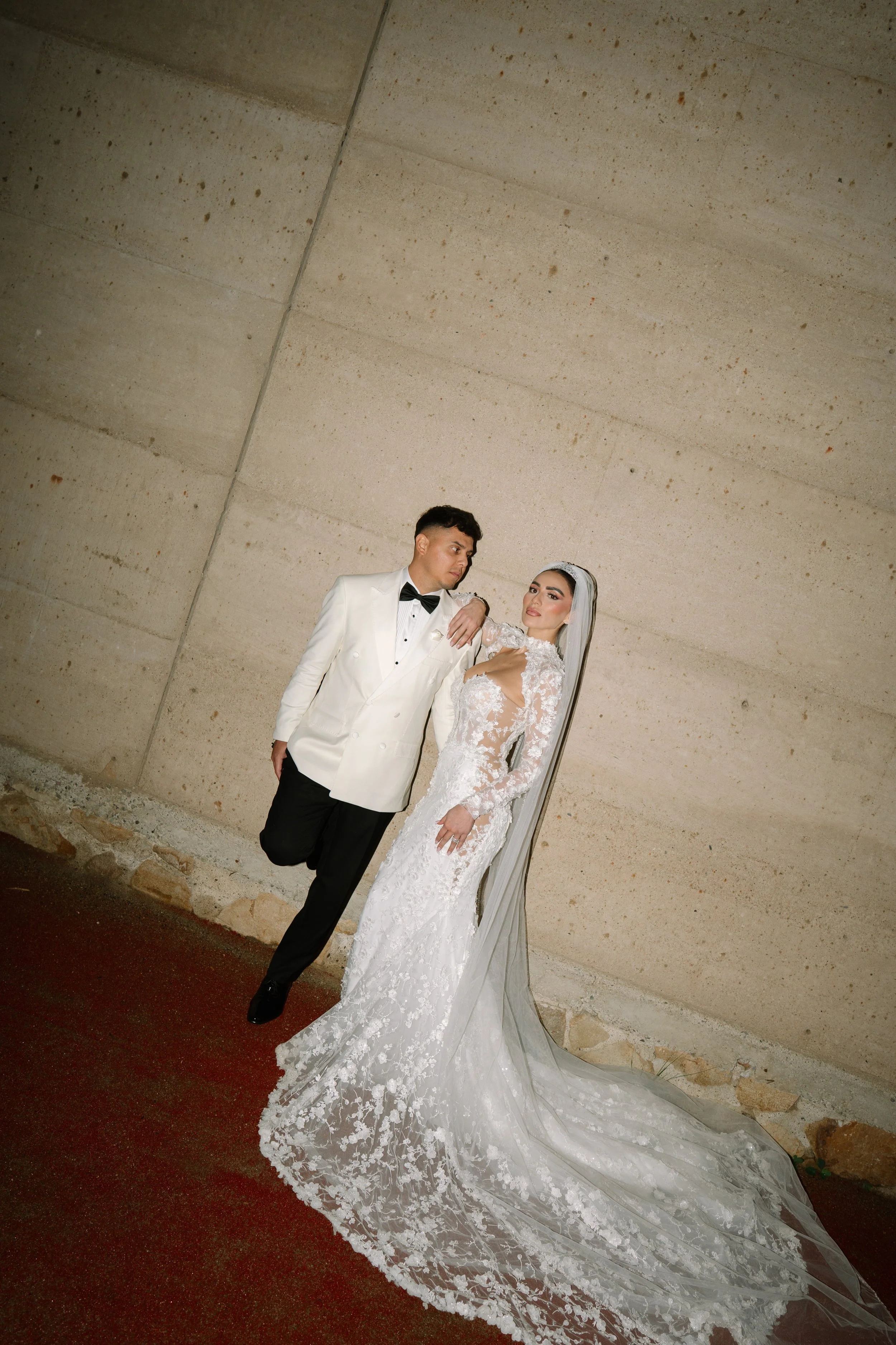 A bride and groom in wedding attire posing against a plain beige wall. The bride is in a white lace wedding gown with a long train and veil, and the groom is in a white tuxedo jacket with black pants and a black bow tie. Wedding in Valle de Guadalupe