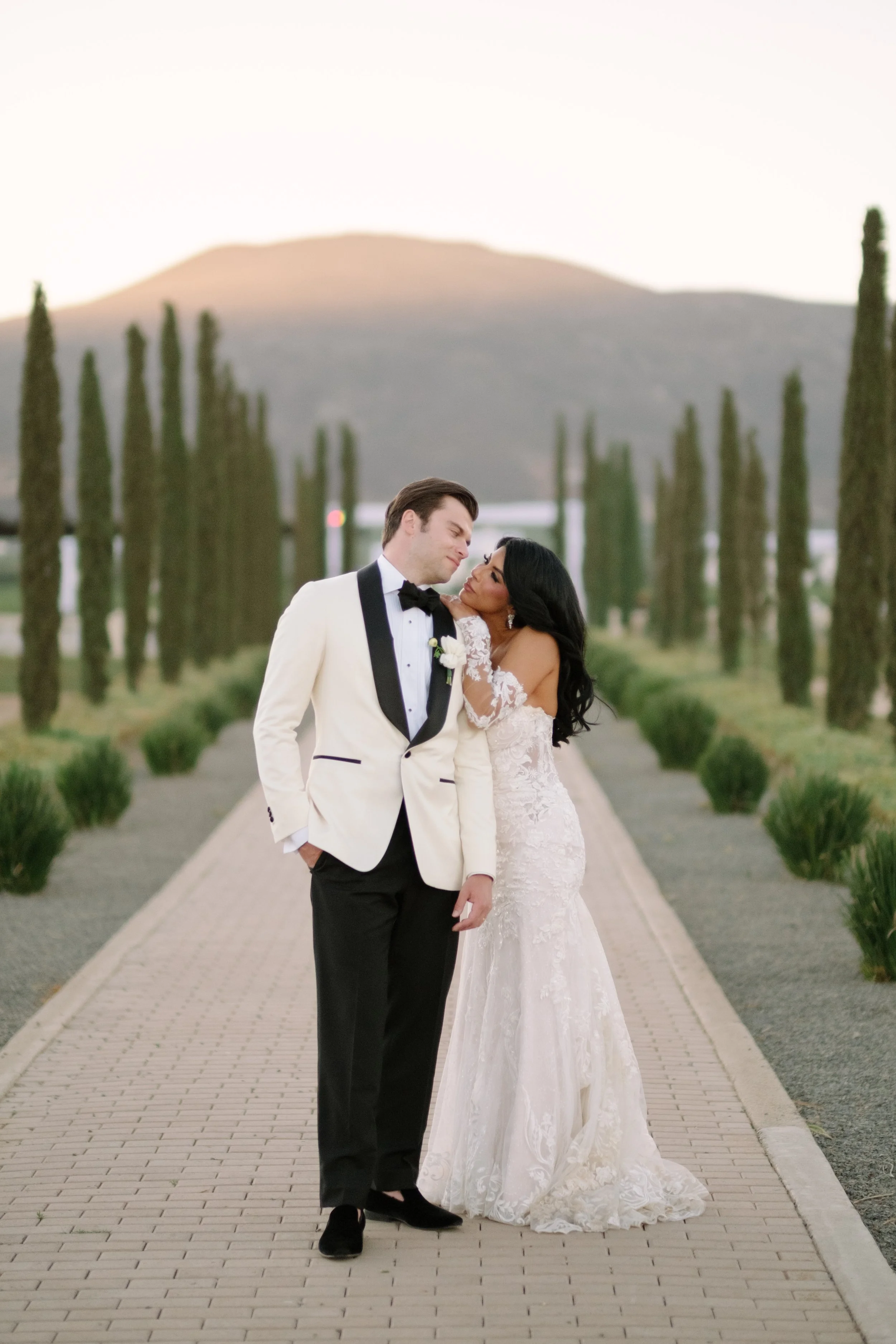 A bride and groom standing on a pathway lined with tall cypress trees, embracing and smiling at each other outdoors during sunset. Wedding Photography in Valle de Guadalupe, Ensenada, Baja California.