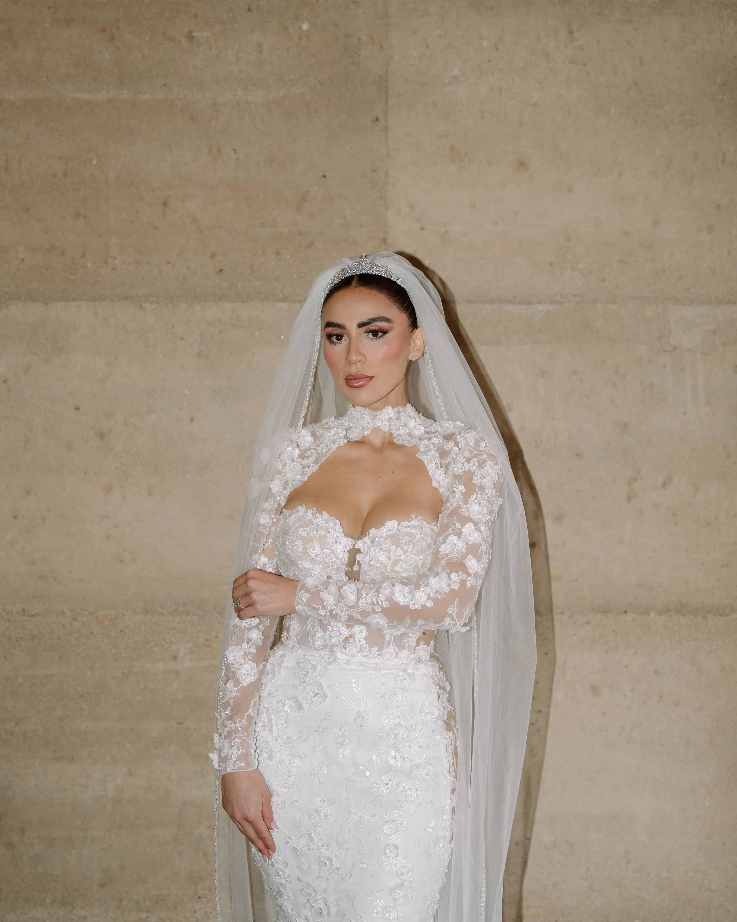 A woman in a white lace wedding dress and veil stands against a beige stone wall. Wedding Photography in Valle de Guadalupe, Ensenada, Baja California.